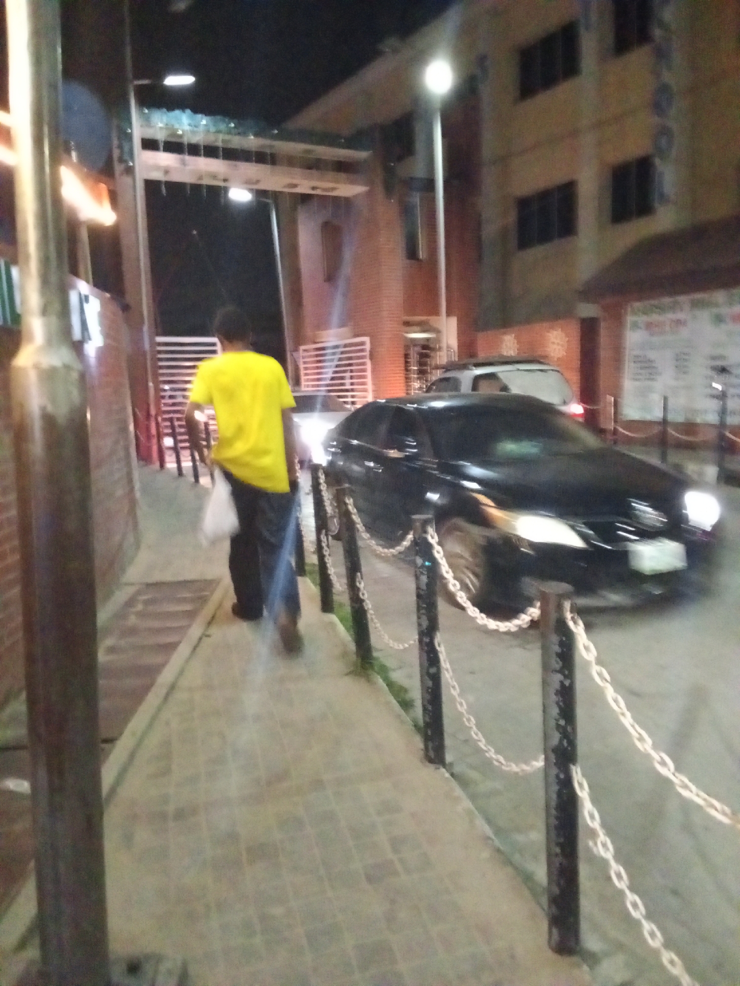 This night-time photograph captures an urban street scene in Ikorodu, Nigeria, featuring a pedestrian and a moving vehicle.
The primary subject is a person walking away from the camera on a paved sidewalk, wearing a bright yellow t-shirt and dark pants. They are carrying a white bag in their left hand. To the left, a large metal pole or pillar stands vertically, partially obscuring the view. The sidewalk has a distinctive checkered pattern.
To the right of the sidewalk, a series of dark metal posts connected by white chains forms a barrier, separating the pedestrian path from what appears to be a driveway or narrow road. A black car, possibly a sedan or SUV, is seen in motion, with its headlights brightly illuminated and showing motion blur, suggesting it is either approaching or driving past. Its front license plate is also visible but illegible. Further in the background, a lighter-colored vehicle, possibly a white SUV, is parked.
The background features a multi-story building with several dark windows and a brownish-tan facade, illuminated by a bright street light on a tall pole. Above the person and extending across the frame is an overhead architectural structure, possibly a gate or archway, fitted with lights. On the far right, part of a signboard with what appears to be illegible text is visible, suggesting a commercial establishment. The overall scene is dimly lit by artificial sources, indicating it is late evening or night. The sky is dark, and there are no discernible weather elements.