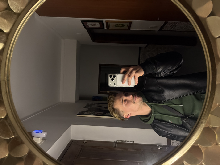 A man takes a mirror selfie. The mirror is round with a gold floral-like design around the edge. The man wears a black leather jacket over a green hoodie. He holds a white iPhone in his right hand. The background shows a living room with wooden furniture, framed pictures on the wall, and a white wall. A small, blue illuminated object sits plugged into an outlet on a wall.