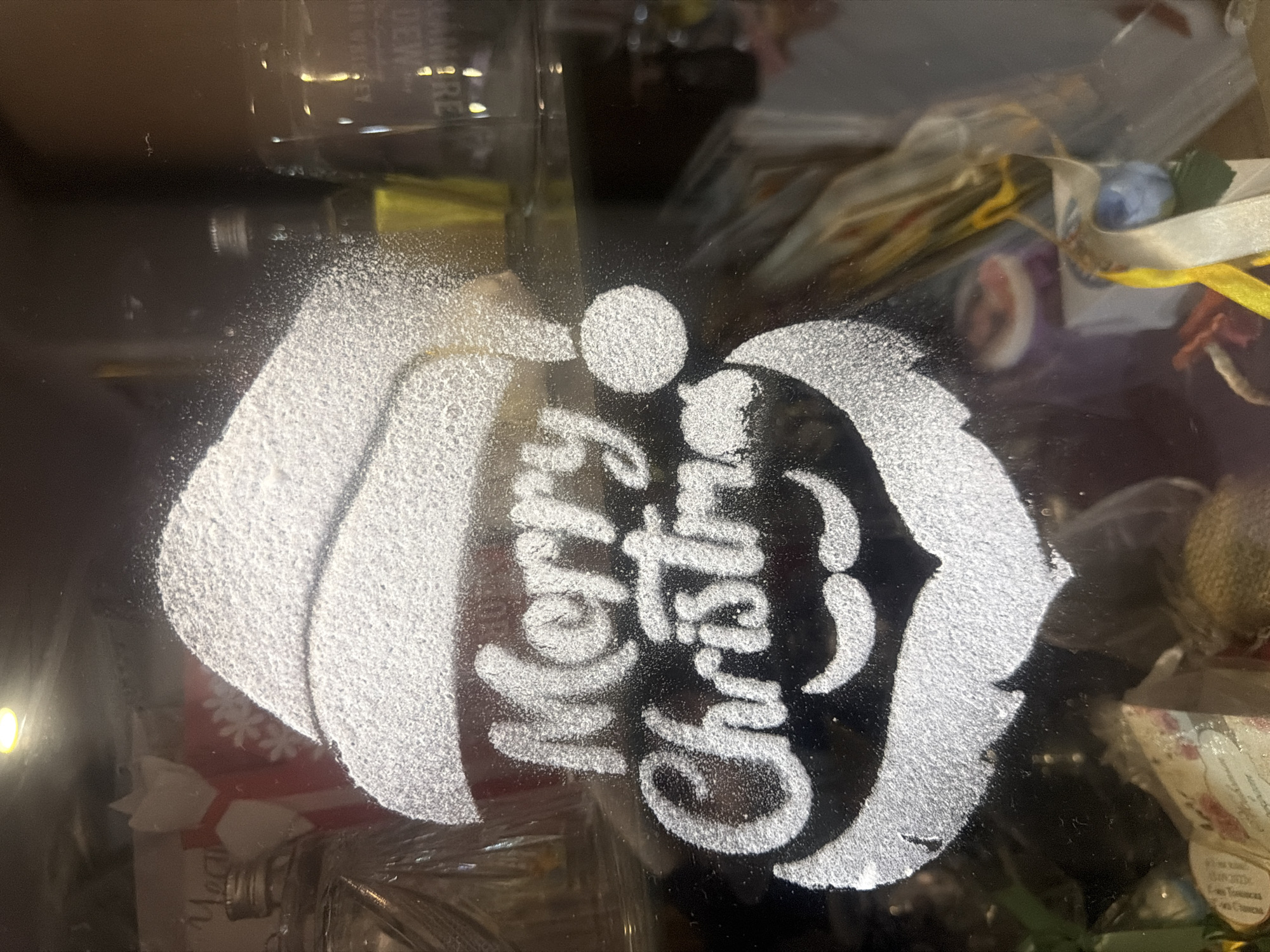 The image shows a drawing of Santa Claus, with the words "Merry Christmas" written on a dark surface, possibly a glass table or countertop. The drawing appears to be made with a powdery substance, possibly powdered sugar or flour. The hat, beard, and lettering are prominent features of the drawing. In the background, there are blurred objects, including what seems to be a bottle and other miscellaneous items, suggesting that the drawing is in a domestic setting. The city and other contextual information about the location are not discernible from the image.