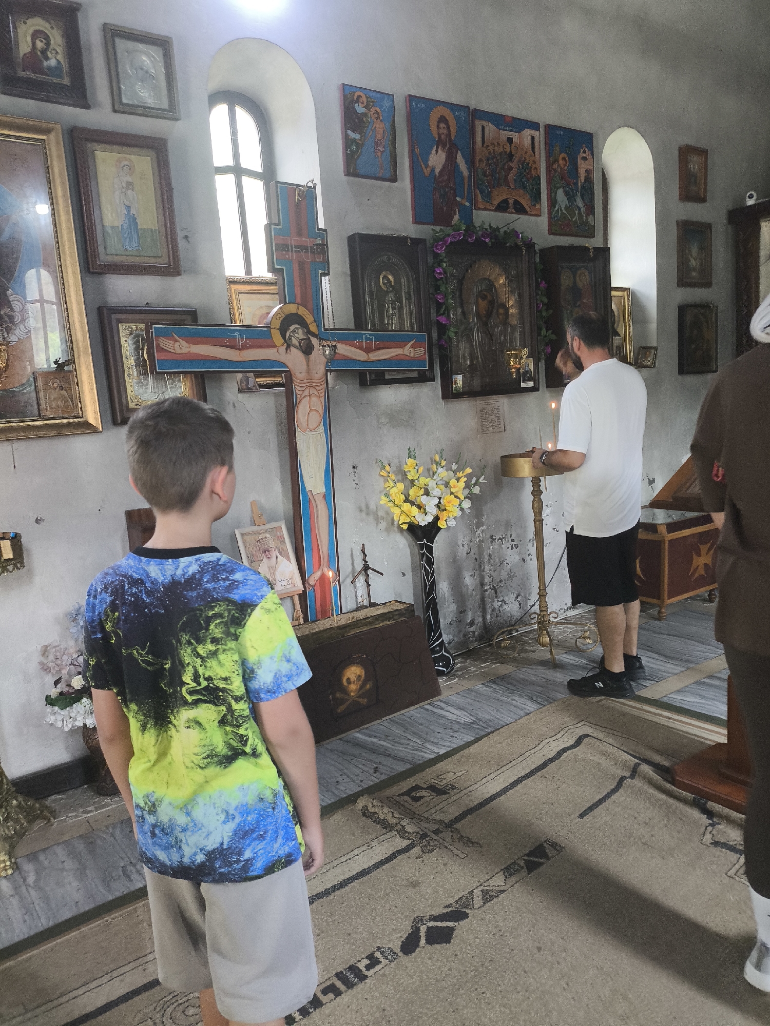 In the interior of a church in Unknown, Unknown, a young boy stands with his back to the camera, facing a large wooden crucifix adorned with religious iconography. He is wearing a t-shirt with a multi-colored abstract design and light-colored shorts. To his right, a man in a white t-shirt and black shorts stands before a tall golden stand with a lit candle, also facing the crucifix. The room features walls covered in religious icons and paintings, illuminated by natural light from a tall arched window. A vase of yellow and white flowers sits on a stand near the crucifix, and a patterned rug covers the floor. Another person, dressed in brown, is partially visible on the far right of the frame.