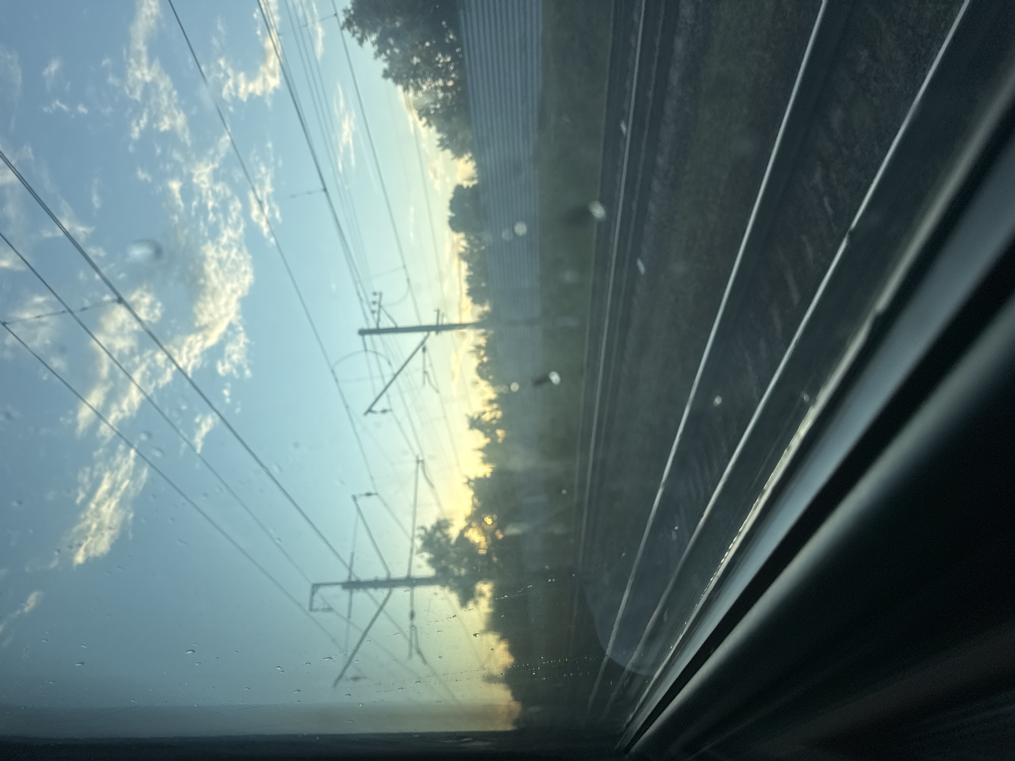 The image captures a view through a train window, looking out at a landscape under a twilight sky. The sky is a gradient of blue and yellow, with scattered white clouds. Overhead power lines for the train are visible, supported by tall poles. Trees line the horizon, interspersed with a sound barrier along the train tracks. The train tracks themselves are in the foreground, stretching into the distance. The window has several water droplets on its surface, some appearing as small circles or irregular shapes. The bottom left of the image shows a dark, metallic structure, likely part of the train's interior or the window frame. The scene appears to be located in or near Malakhovka, Russia, given the presence of typical Russian railway infrastructure and vegetation. There are no people or animals visible in the image.