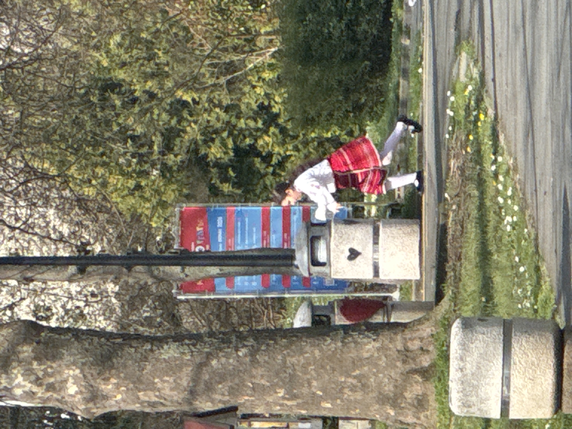 A woman wearing a white shirt and a red plaid skirt is bent over, seemingly interacting with a trash bin located next to a street. The bin is grey with a black heart symbol on its side. Nearby, a sign with blue and red coloring is visible. The environment includes a paved area with vegetation alongside it. A large tree trunk is in the foreground. Based on the sign's lettering, the location is identified as Burgas, Bulgaria.
