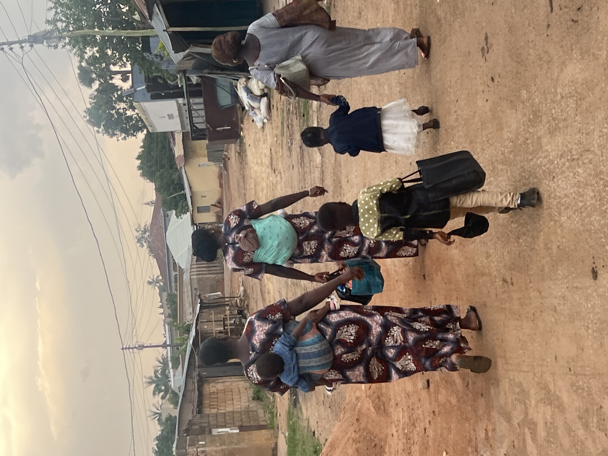 The image depicts a group of individuals walking along a dirt road in what appears to be a residential area of Gura Top, Nigeria. Several women, some wearing patterned dresses, are present. One woman carries a child on her back. Another woman is holding the hand of a young girl in a white skirt. Another person is carrying a black bag. The surrounding environment consists of simple structures, possibly homes, with vegetation and overhead wires visible. The scene appears to be captured during daylight hours.