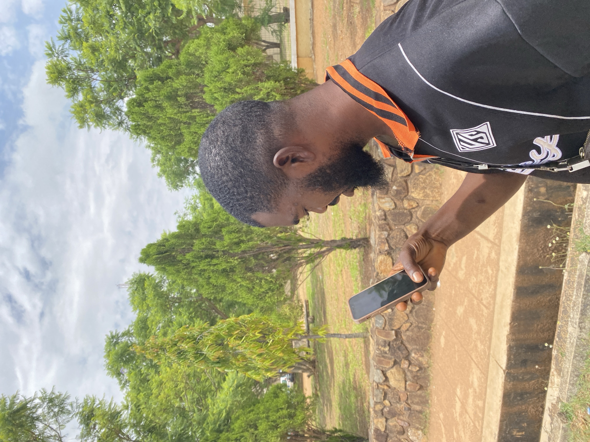A man with dark skin and a beard stands outdoors, looking down at a cell phone held in his right hand. He is wearing a black shirt with orange and white detailing. The background consists of green trees and a partly cloudy sky. The ground appears to be a park-like setting, with a stone structure visible near the bottom right. The image is oriented vertically. The city of Gwong, Nigeria is not definitively identifiable in this image, but the environment suggests an outdoor setting.