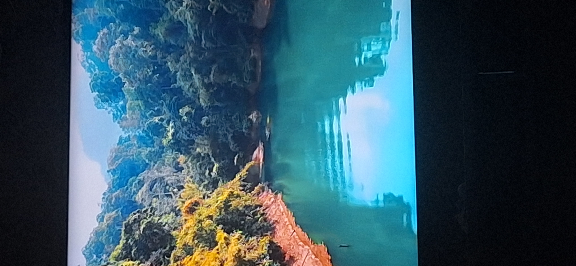 The image shows a serene natural landscape. A body of turquoise water fills the lower half of the frame, reflecting the bright sky and surrounding vegetation. On the left side, a reddish-brown bank slopes down to the water, with some green and yellow foliage growing on it. The upper portion of the image is dominated by dense green trees covering rolling hills or mountains, with a hazy sky above. There are no people or man-made structures visible in the scene. The location in Bila Tserkva, Ukraine, is not depicted in this image.