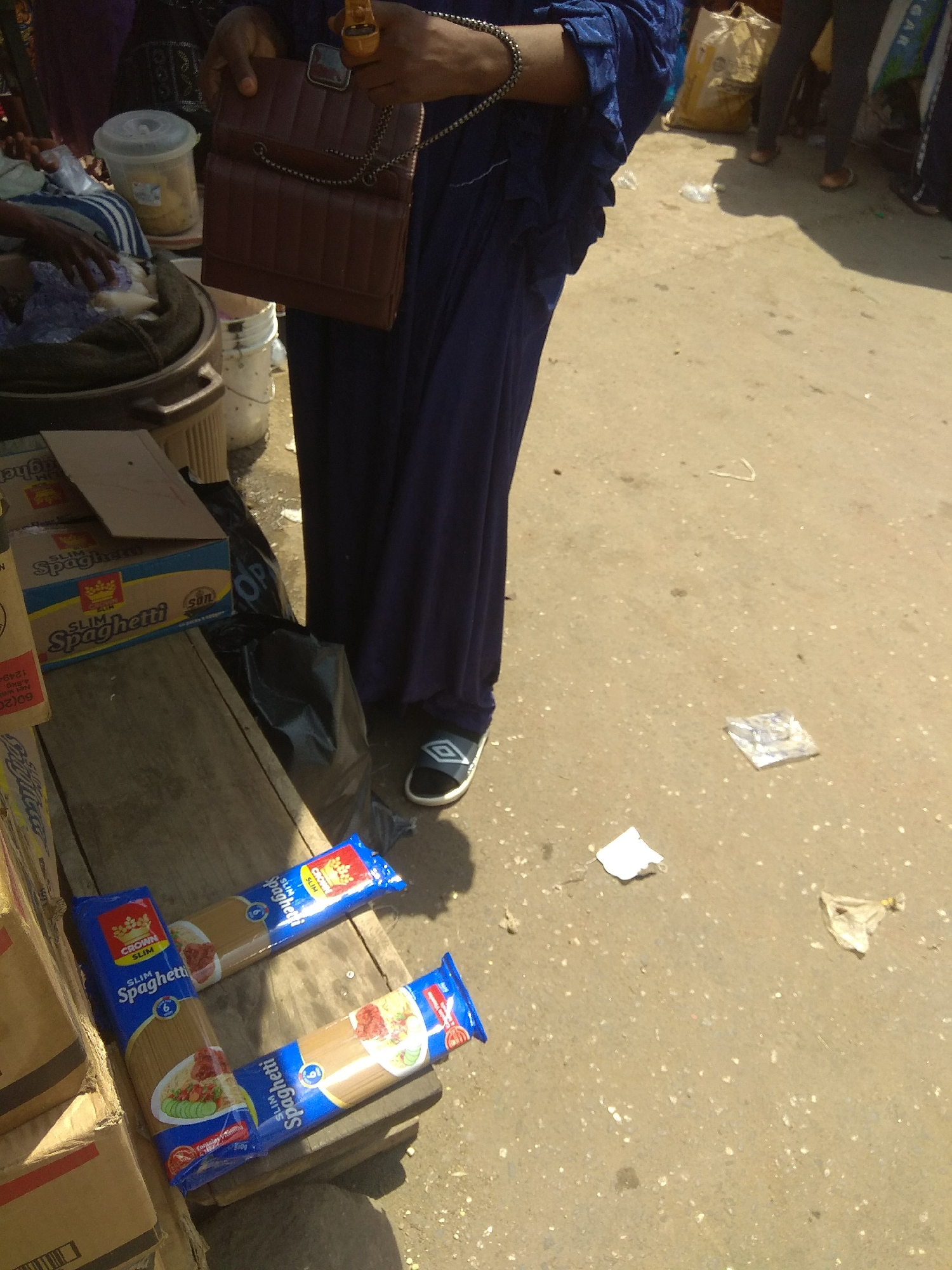 The image shows a close-up view of a person, likely a woman based on the attire and handbag, standing in what appears to be an outdoor market setting. The person is wearing a long, dark blue garment and holding a brown, quilted handbag with a chain strap. Their feet are visible, wearing dark shoes with a white diamond pattern. In the foreground, there are two packages of "CROWN SLIM Spaghetti" laid out on a wooden surface, with some cardboard boxes to the left. The background is somewhat blurred, but shows other people and market goods, including what looks like a large yellow bag and a white bucket with a lid. The ground is paved with rough concrete, and there are various pieces of litter scattered around, including plastic wrappers and paper. The lighting suggests it is daytime, likely sunny, casting shadows on the ground. The scene conveys a sense of everyday commerce in a bustling outdoor market, possibly in Jalingo, Nigeria, given the location context. The focus is on the spaghetti products and the person's attire and accessories.