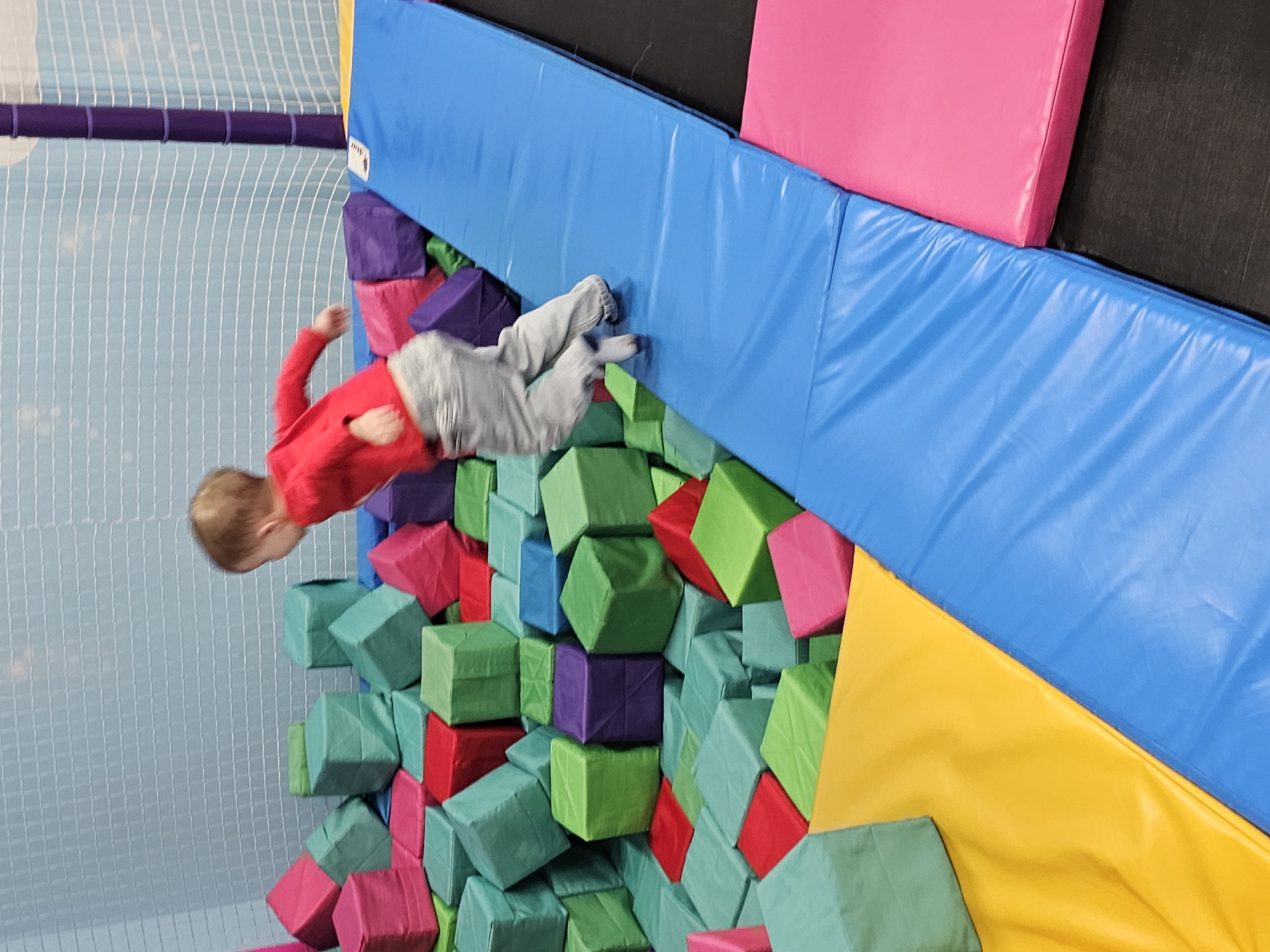 A child is in mid-air, falling toward a pit filled with colorful foam blocks (red, green, purple, blue) at a trampoline park. The pit is bordered by padded surfaces in yellow, blue, and pink. A netting structure is visible in the background. The location is Khimki, Russia, indicated by a sign (partial view) in Russian near the upper left.