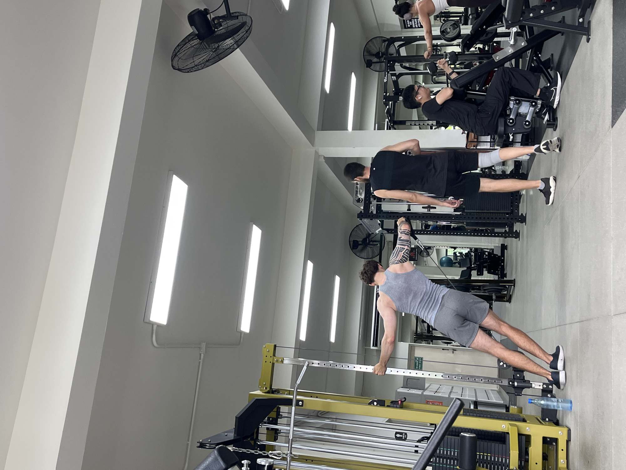 The image depicts the interior of a gym in Chiang Mai City Municipality, Thailand. The space is well-lit with fluorescent ceiling lights and features various weightlifting and exercise equipment. In the foreground, a man with extensive arm tattoos is performing a cable crossover exercise, leaning back and pulling the handles towards his chest. He is wearing a grey tank top and grey shorts. To his right, another man in a black tank top and black shorts, with a knee brace on his left leg, stands facing away from the camera. Further back, a person wearing glasses and a black t-shirt is seated on a weight machine, appearing to be lifting weights. To the far right, a woman with her hair in a bun, wearing a white top, is also engaged in a workout. The floor is a light grey tile. A blue water bottle rests on the floor near the man doing cable crossovers. Fans are mounted on the ceiling. A fire exit sign with Thai text is visible on a wall in the background.