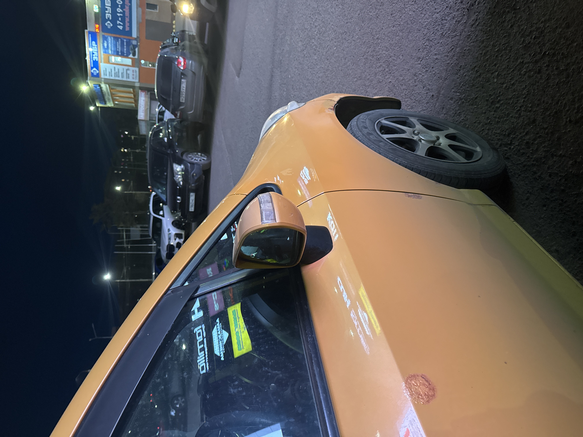 The image shows an orange car parked on the side of a road in Izhevsk, Russia. A tire is visible on the right side of the frame. The car has stickers on its windows, and a side mirror. In the background, there are several other cars and a building with signage in Russian. The streetlights are on, suggesting it is either evening or night. The image appears to be taken at street level, with a close-up view of the orange car.