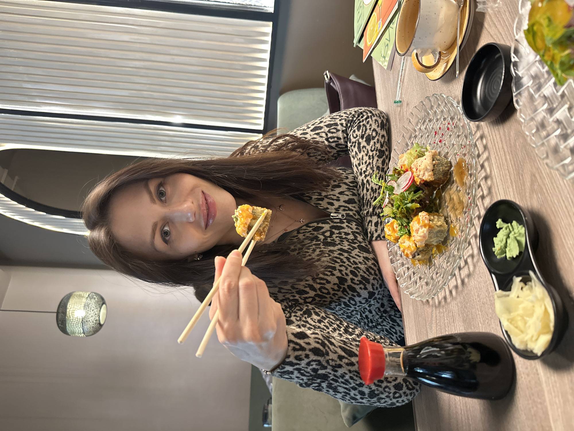 The image captures a woman seated at a table in what appears to be a restaurant, enjoying a meal. She has long, dark hair and is looking directly at the camera with a subtle smile. She is wearing a leopard print top or dress with a zippered front. In her right hand, she holds a pair of chopsticks, picking up a piece of food, possibly a sushi roll.
On the wooden table in front of her, a clear, patterned glass plate holds several pieces of deep-fried or tempura-style sushi rolls, garnished with green salad leaves and radish slices. Next to this, a black, two-compartment dish contains bright green wasabi and thinly sliced pickled ginger. A dark bottle with a red cap, likely soy sauce, is also on the table. Further back, an empty cup and saucer are visible, along with another small, empty black bowl.
The background features a window with horizontal blinds, allowing natural light into the space. A modern, metallic hanging light fixture can be seen on the left. On the far side of the table, upright menus or flyers with colorful designs are present, though any text on them is illegible. The setting suggests an indoor dining experience, likely at a casual restaurant or cafe in Murino, Russia. The overall atmosphere appears relaxed and pleasant.