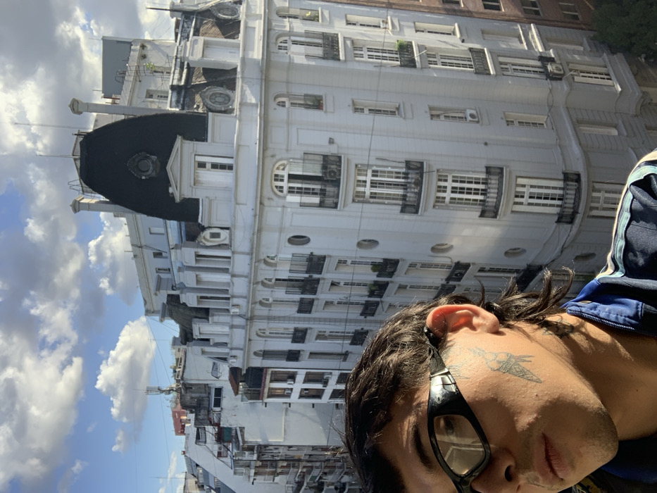 A building with white facade and multiple windows stands tall under a partially cloudy sky. A person with sunglasses is in the foreground. They have dark hair and a tattoo on their cheek. The person is wearing a blue jacket with white stripes on the sleeve. The image is angled, giving a tilted perspective of the building and the person.