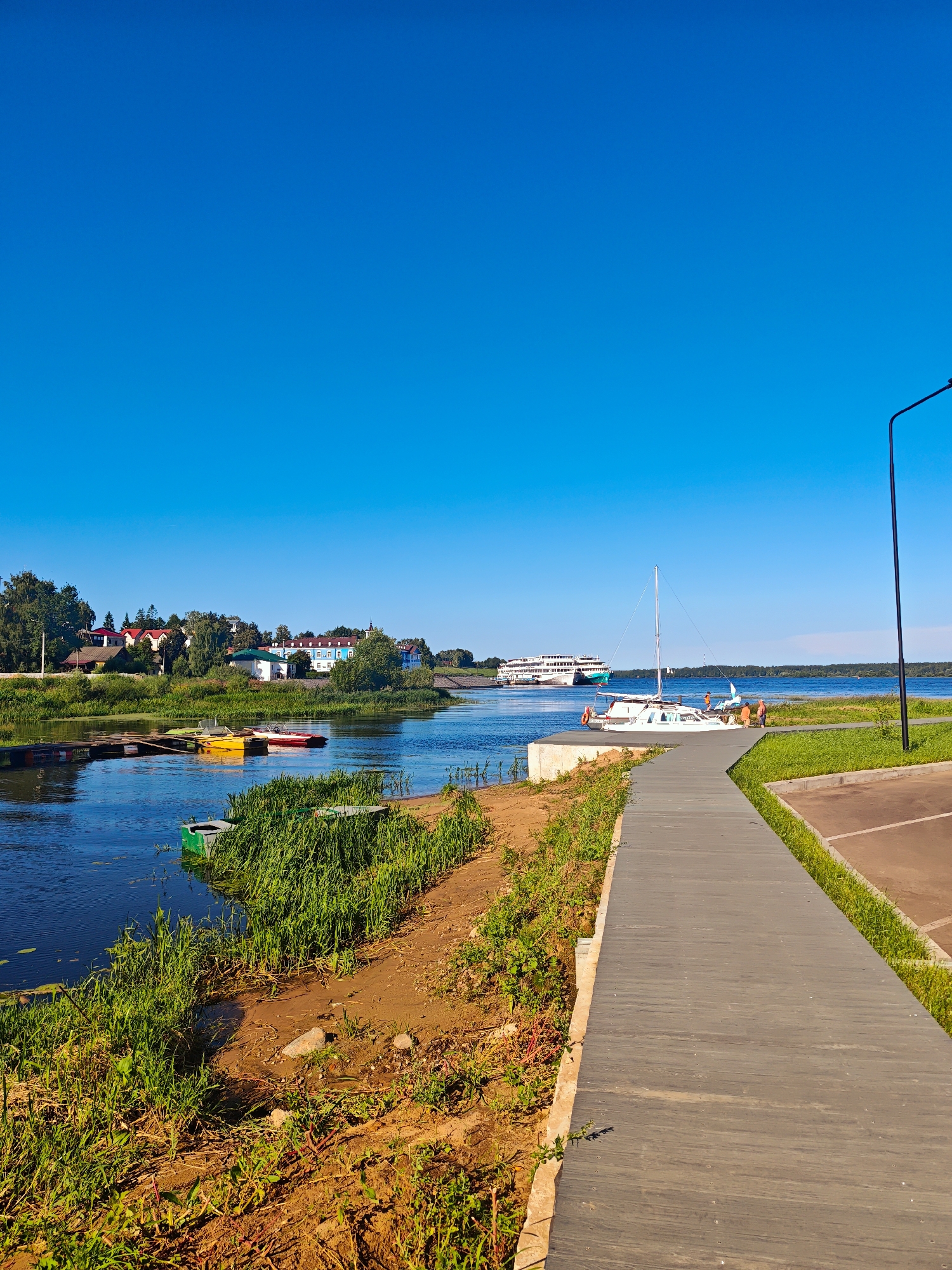 The image captures a scene along the Volga River in Myshkin, Russia, on a clear, sunny day. A wooden boardwalk runs diagonally from the foreground to the middle right of the frame. To the left of the boardwalk, the river is visible, with several small boats docked near a wooden pier. In the distance, on the opposite bank, is a town with buildings and trees. Further out on the river, a large passenger ship is sailing. A white sailboat is moored near the boardwalk, with a few people on board or nearby. The boardwalk is bordered by a grassy area and what appears to be a parking lot. A lamppost stands to the far right of the image. The sky is a vibrant, cloudless blue.