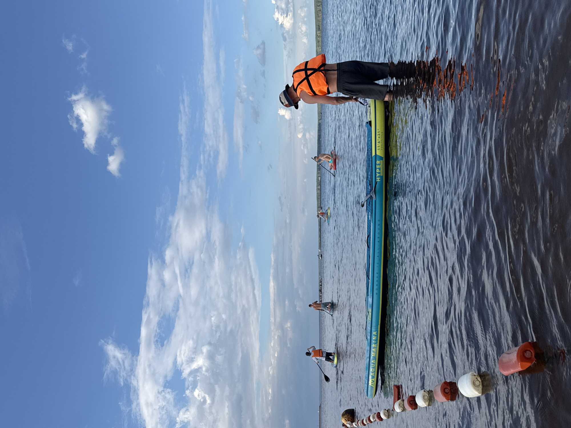 A group of people are stand-up paddleboarding on a calm body of water under a bright blue sky with scattered white clouds. The sun appears to be high in the sky, suggesting it is daytime. The water is dark and reflects the sky, with some ripples indicating gentle movement.
In the foreground, a man wearing an orange life vest and a baseball cap is standing on a paddleboard, holding a paddle. Another paddleboard, a blue and yellow Aqua Marina brand with the text "WONDER IS ALL AROUND" and "AQUA MARINA" visible, is partially in the frame. The man's reflection is visible in the water, along with the orange reflection from his life vest.
Further in the background, several other individuals are also paddleboarding. One person in a light-colored bathing suit is visible on a paddleboard, as is another person wearing an orange life vest. The scene suggests a recreational activity taking place at a lake or bay.
In the distance, a narrow strip of land with some greenery is visible, possibly indicating the shoreline. A line of buoys floats on the water in the foreground, separating a section of the water. The overall mood is relaxed and enjoyable, indicative of a pleasant day spent outdoors. The provided location context is Chaikovsky, Russia.