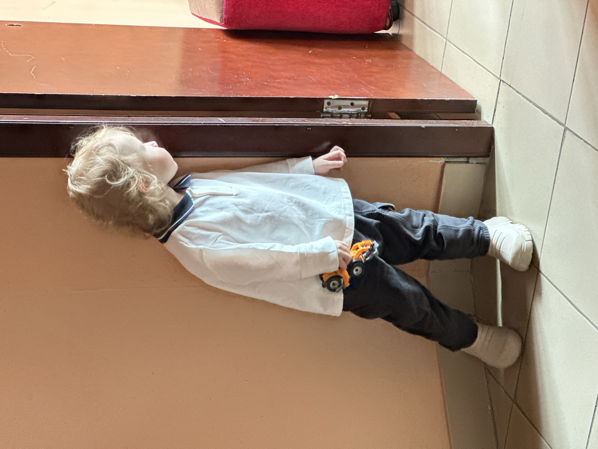 A young child is lying on a beige floor, possibly tile or linoleum, adjacent to a set of dark brown stairs or a low platform with a red object atop it. The child is wearing a white long-sleeved shirt with a dark blue collar, dark gray pants, and white shoes. They are holding a small orange toy truck. The location appears to be indoors, near a tiled wall. This location cannot be determined to be located in Votkinsk, Russia.