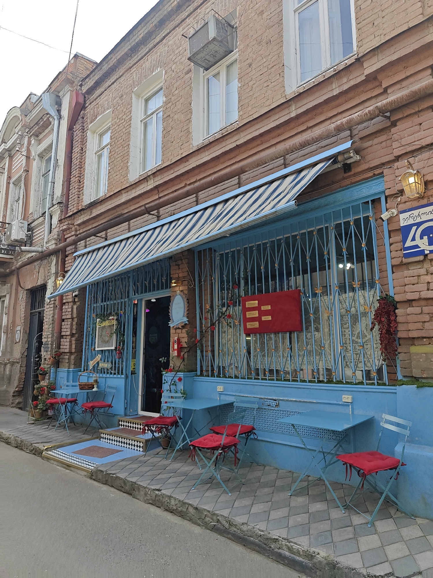 This media file captures a charming, light blue shopfront on a narrow street in Tbilisi, Georgia, with an old, weathered brick building rising above it. The scene suggests a cafe or small eatery, set up with outdoor seating.
The shop's facade is painted a distinctive light blue, featuring intricate security grates over its large windows and entrance door. A blue and white striped awning extends over the outdoor seating area. In front of the shop, several light blue metal tables and chairs with bright red cushions are arranged on the paved sidewalk, inviting passersby. Red roses, possibly artificial, adorn the grates and an oval sign near the entrance, adding a splash of color. A small woven basket with an "OPEN" sign hangs near the doorway.
Above the shop, the building reveals a worn reddish-brown brick exterior with white-framed windows. A rusty metal pipe runs horizontally across the brickwork, while an older, adjacent building shows peeling paint and a visible drainpipe. An old air conditioning unit is mounted on the upper left side of the building.
Notable details include a warm-glowing wall lantern and a CCTV camera mounted above a blue sign that features Georgian script and "4G". Inside the shop window, a red rectangular board with multiple lines of Georgian text is displayed. The sidewalk in front of the shop is paved with square and diamond-patterned tiles, and a small step-up entrance to the shop is marked with black and white checkered tiles. The sky appears overcast, suggesting it's daytime but without direct sunlight. There are no people visible, giving the scene a serene and quiet atmosphere.