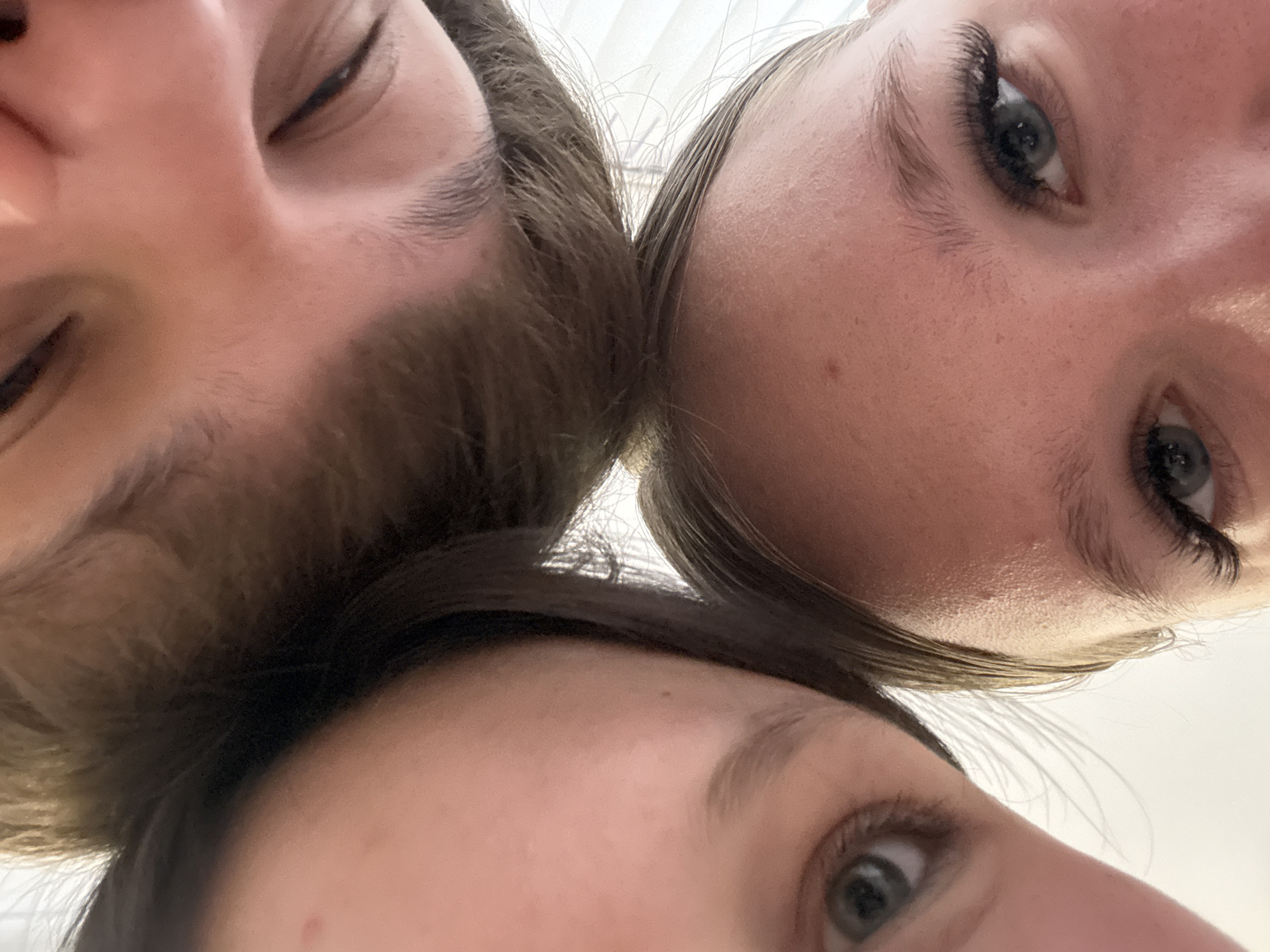 Three individuals are closely positioned, with their faces in close proximity to each other. The faces fill the frame. The individuals are looking down at the camera. The angle is such that the top of each head touches the frame. The focus appears to be mainly on the eyes. The environment is undefined but appears to be indoors due to the presence of artificial light coming from what looks to be a light fixture. There is no specific information about the location being Chaikovsky, Russia.