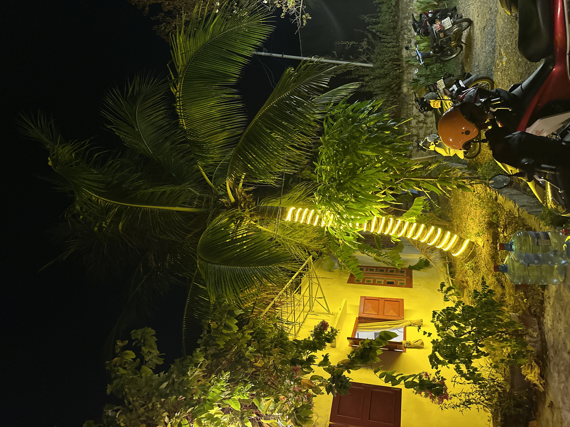 The image depicts a nighttime scene in Bac Nha Trang Ward, Vietnam. A yellow building with red-framed windows and doors is illuminated by artificial light, including a string of lights wrapped around a palm tree. Several motorcycles are parked along a wall in the background. A stack of water bottles is visible near the base of the building. Vegetation surrounds the building, partially obscuring it. The overall impression is that of a brightly lit building surrounded by darkness and greenery.