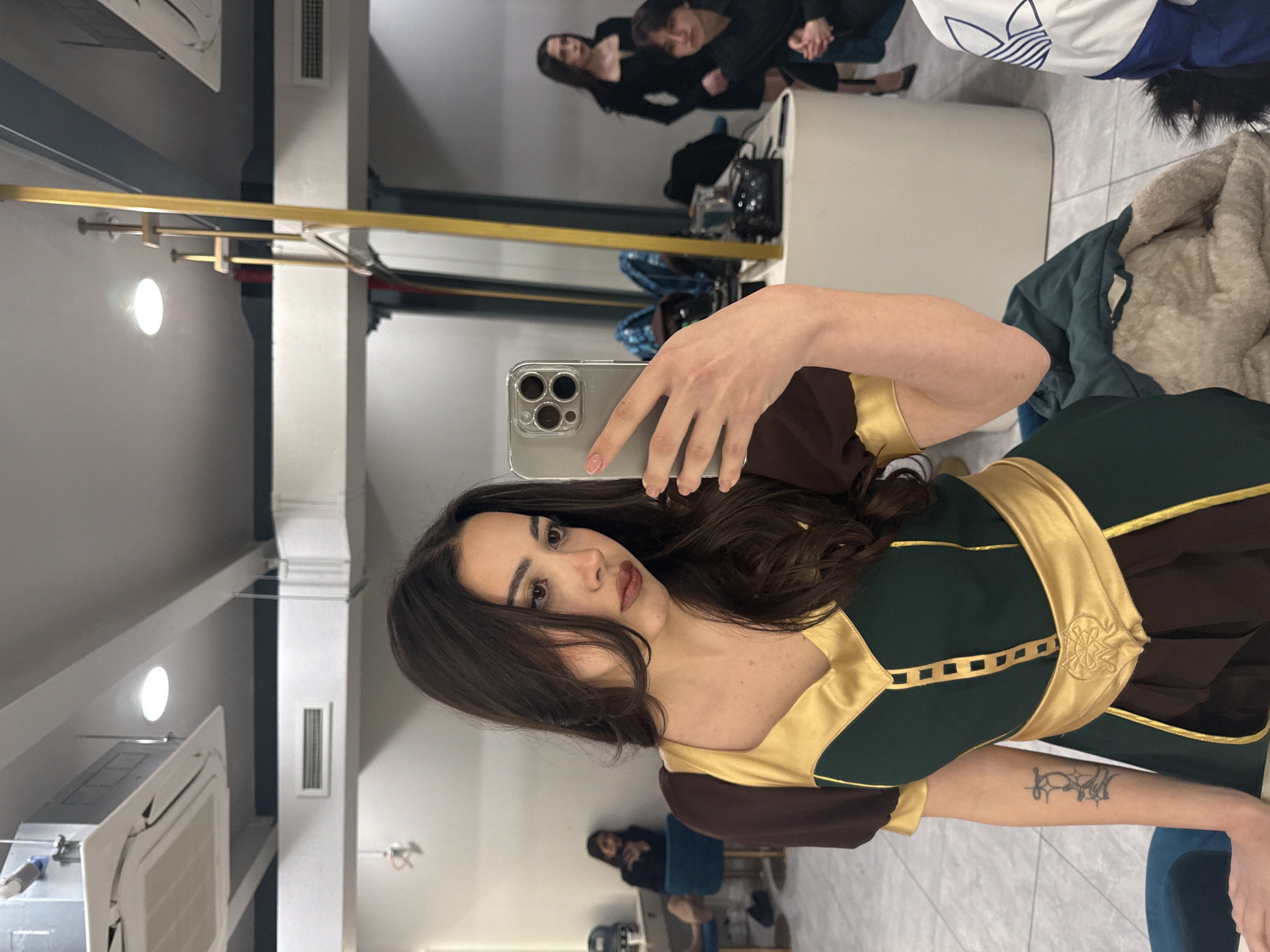 A young woman takes a selfie in a mirror. She is wearing a brown and green costume with gold trim. The woman has long, dark hair and brown eyes. The selfie is taken with an iPhone.
In the mirror reflection, other women are visible. The location is Yerevan, Armenia.