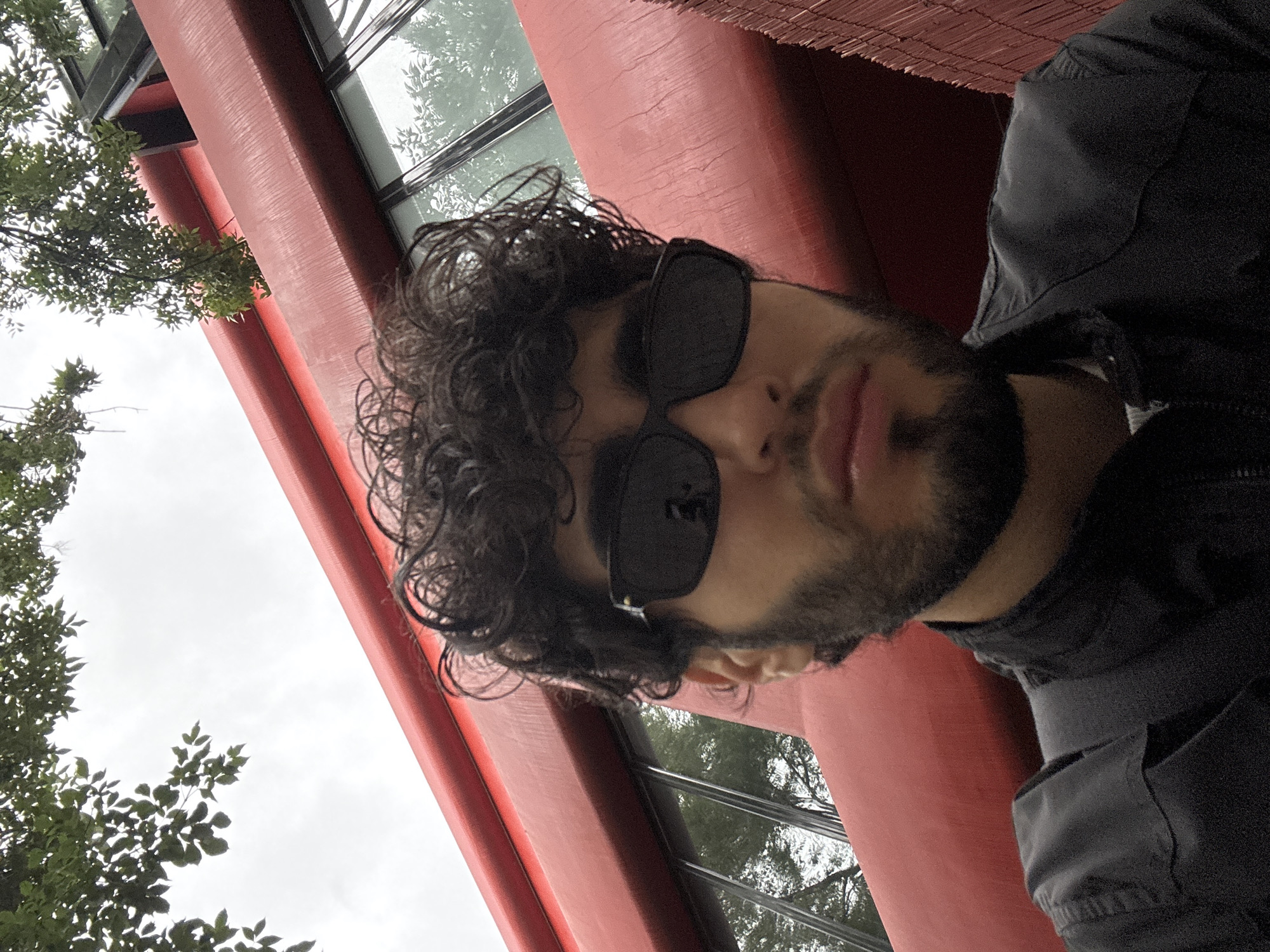 The image is a selfie of a young man wearing sunglasses and a dark jacket, taken from a low angle. He has curly dark hair and a beard. Behind him, a distinctive red building with large windows is visible, with green tree branches overhead against a cloudy sky. The building's architectural style suggests it could be part of a cultural or educational institution. Based on the architectural design and prominent red color, the building is identifiable as the National Gallery of Armenia, located in Yerevan, Armenia.