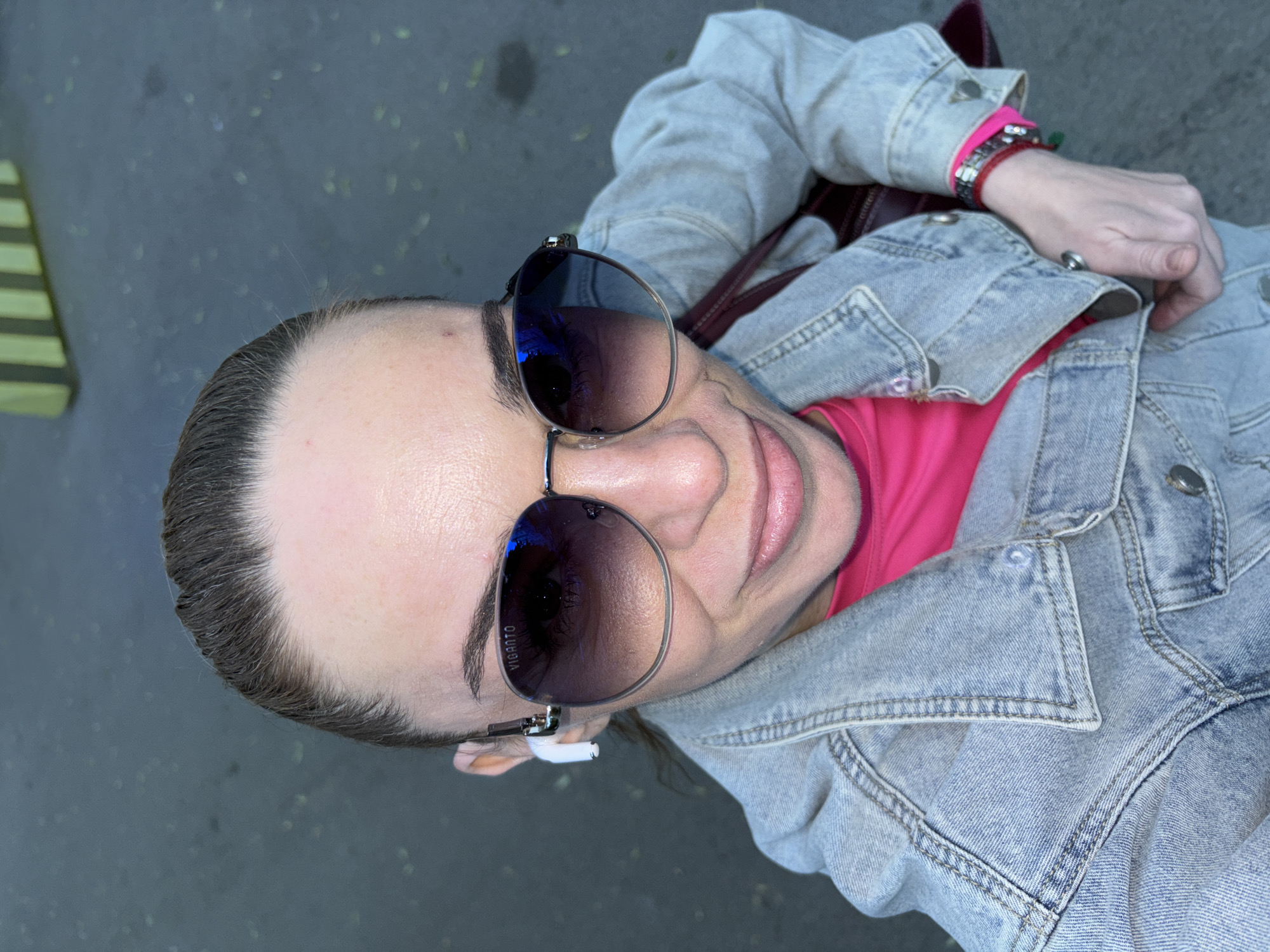 A woman is captured in a high-angle, close-up selfie in Sinyavino, Russia. She is wearing large, aviator-style sunglasses with a gradient tint and a silver frame, which display a reflection of the sky. Her hair is pulled back tightly, revealing a single white wireless earbud in her left ear. She has on a bright pink long-sleeved shirt underneath a light-wash denim jacket. A maroon handbag strap is visible across her shoulder. Her right hand is resting on her jacket, and her left wrist is adorned with bracelets, including a bright pink band. The background is out of focus, showing a grayish, textured surface, possibly pavement, with some scattered green leaves.