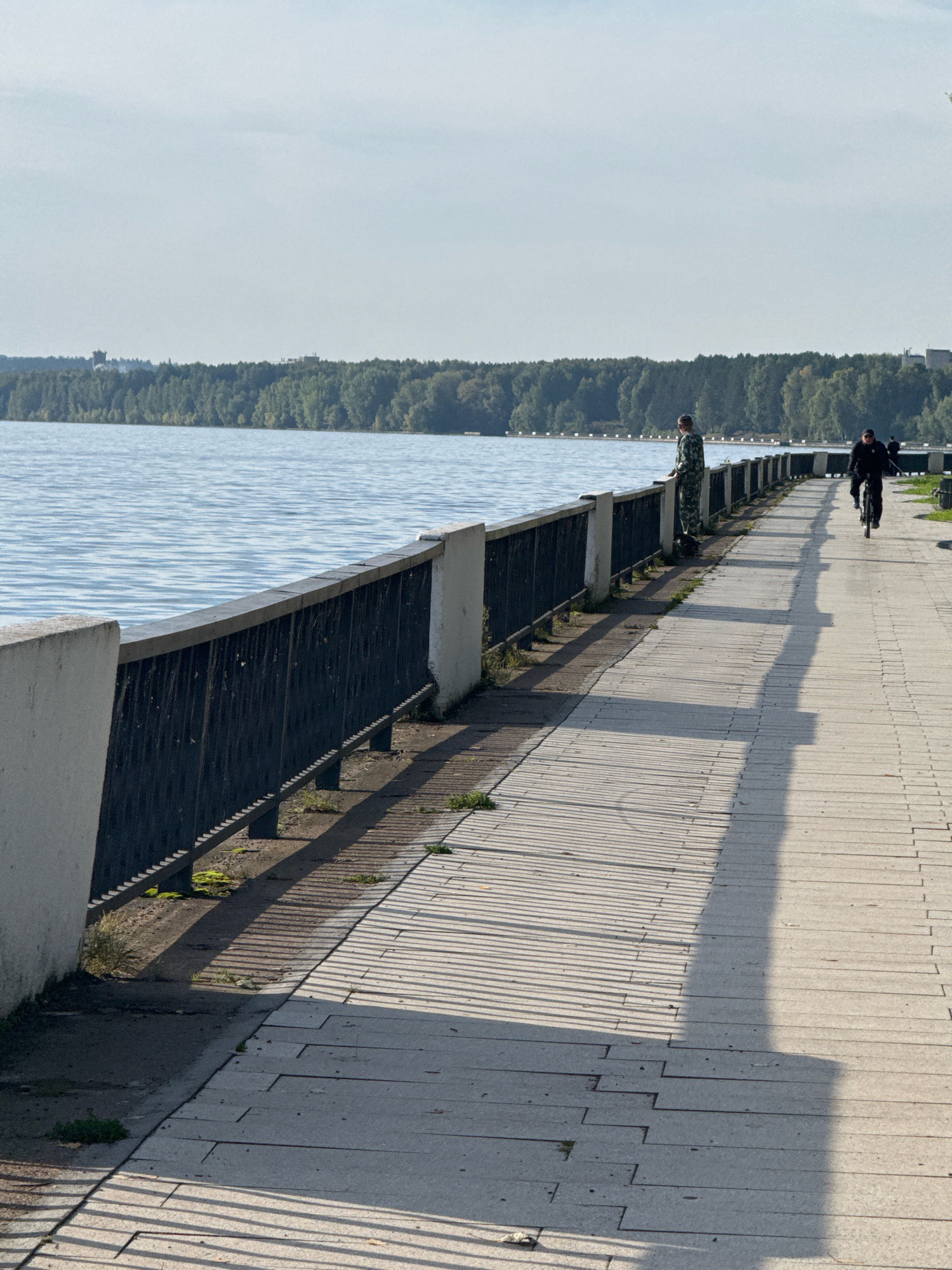 This image captures a view of the Kama River embankment in Chaikovsky, Russia, on a sunny day. The embankment is paved with light-colored rectangular stones, creating a textured pathway. A dark metal fence with white concrete posts runs along the edge of the river, separating the walkway from the water. The Kama River is a tranquil blue, reflecting the clear sky.
In the foreground, a person in camouflage clothing stands near the fence, looking out at the river. Further down the embankment, another person is riding a bicycle. The sun casts long, distinct shadows from the fence onto the paved walkway. In the background, a dense line of green trees forms a natural border along the far bank of the river, with some structures visible in the distance. The overall scene suggests a peaceful, everyday moment on a public promenade.