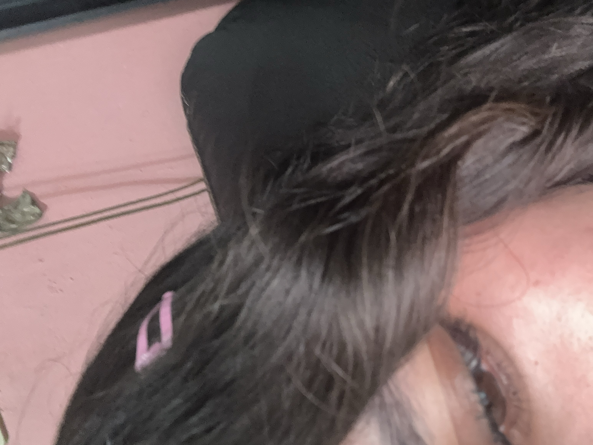 A close-up photograph shows a portion of a person's face. The person has dark brown hair with a pink bobby pin visible. The eye is visible, with dark lashes. The surrounding skin is fair. A black head covering is visible on top of the head. The background is pink. There are no discernible indicators of the location being San Cristóbal, Mexico.