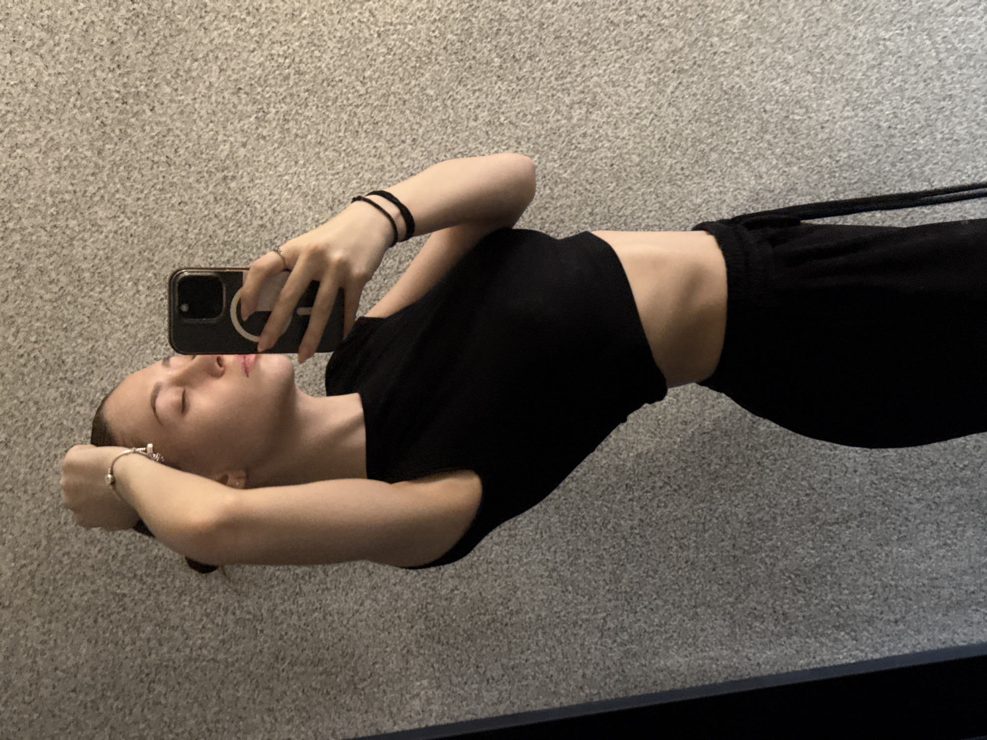 A woman is lying on a gray carpet, taking a selfie with her phone. She is wearing a black crop top and black sweatpants. She has a bracelet on her left wrist and black elastic bands on her right wrist. Her eyes are closed. The bottom of a mirror is visible in the lower portion of the image. The location is unknown, there is no indication that the image was taken in Sofia, Bulgaria.