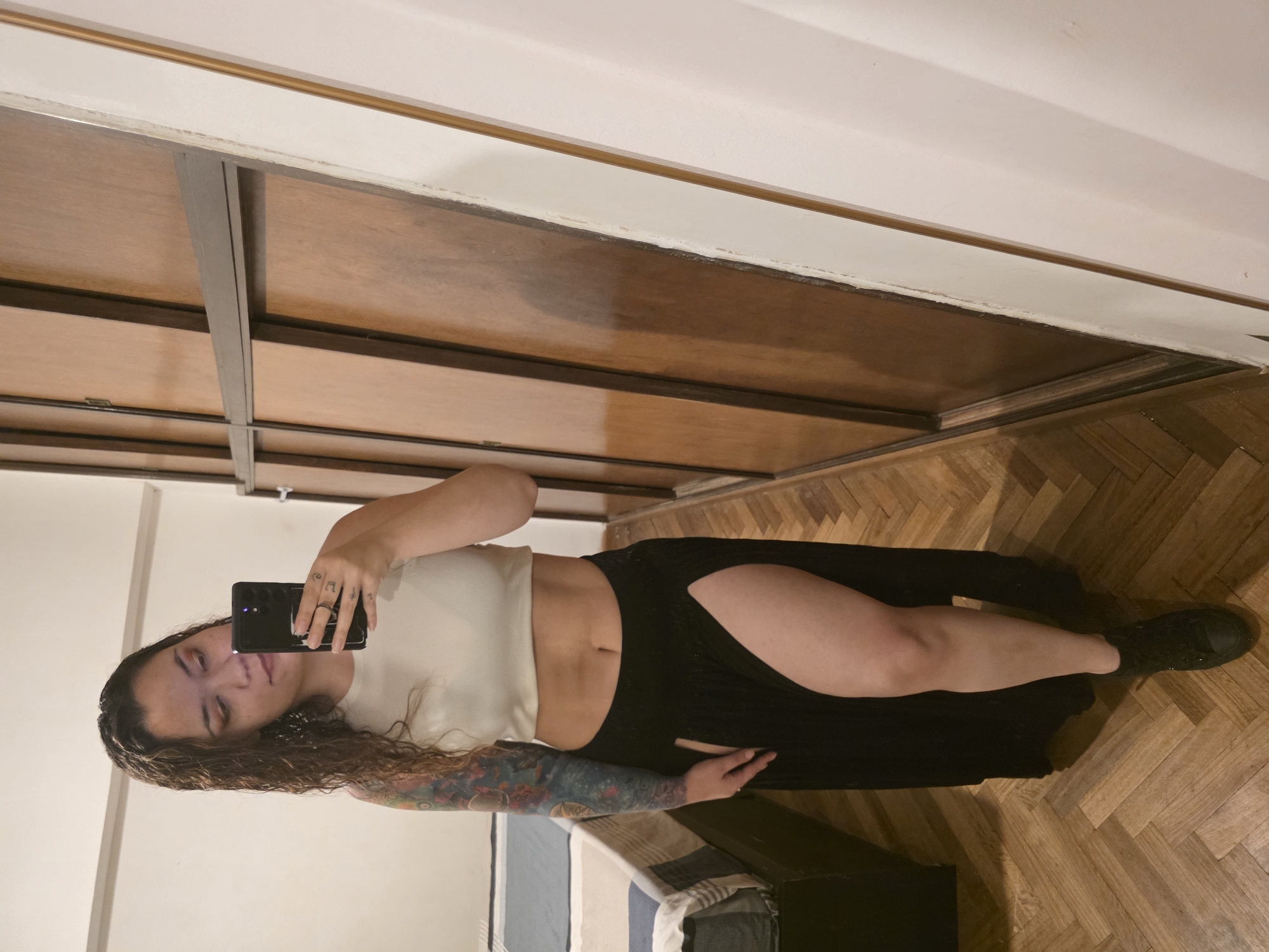A single female subject is depicted standing and taking a mirror selfie in an interior residential space, likely a bedroom or dressing area, in an apartment in Buenos Aires, Argentina. This inference is drawn from the common herringbone parquet flooring and the style of the built-in wooden wardrobe.
The subject is a light-skinned female with long, wavy brown hair, parted on the right. She is looking down at a black smartphone held in her right hand, which is raised to eye level. Her left arm is extensively tattooed with a full-sleeve design featuring multiple colors, visible from shoulder to forearm. She is wearing a light cream or off-white sleeveless crop top that exposes her midriff, and a long black skirt with a high slit on the left side, revealing her left thigh and leg. On her feet are black low-top sneakers. A ring is visible on the ring finger of her right hand, and another on her thumb.
The environment includes a prominent floor constructed from wooden parquet laid in a herringbone pattern. To the right of the subject is a large, built-in wooden wardrobe or closet composed of multiple vertical panels, with dark grey horizontal and vertical framing elements. The surface of the wardrobe appears somewhat aged. To the left and behind the subject, a portion of a bed is visible, covered with a duvet featuring horizontal stripes in shades of blue and white, with a darker block at the head of the bed. The walls are predominantly light-colored, likely white, and show minimal detail. The lighting is internal and appears consistent throughout the room.