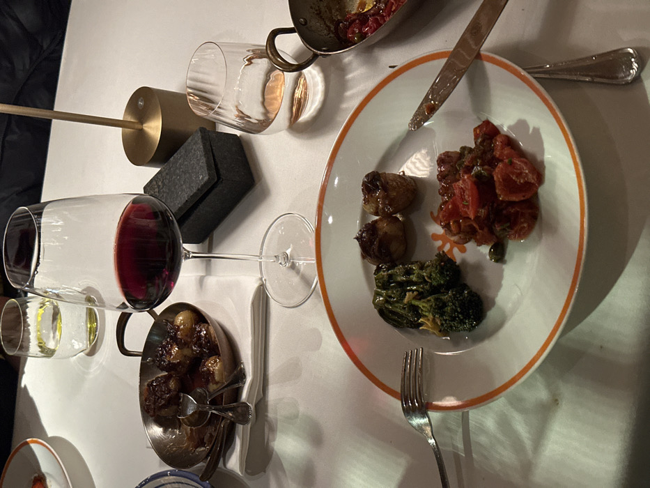 An overhead shot showcases a white tablecloth setting with a plate of roasted potatoes, tomatoes, and broccoli. Adjacent is a small pan filled with similar potatoes and serving utensils. A glass of red wine and a shorter glass are also present, along with a bronze cylinder base with a long pole extending to the left and a dark gray square. A pan with cooked items and a knife are positioned at the top. A fork rests near the plate's bottom.