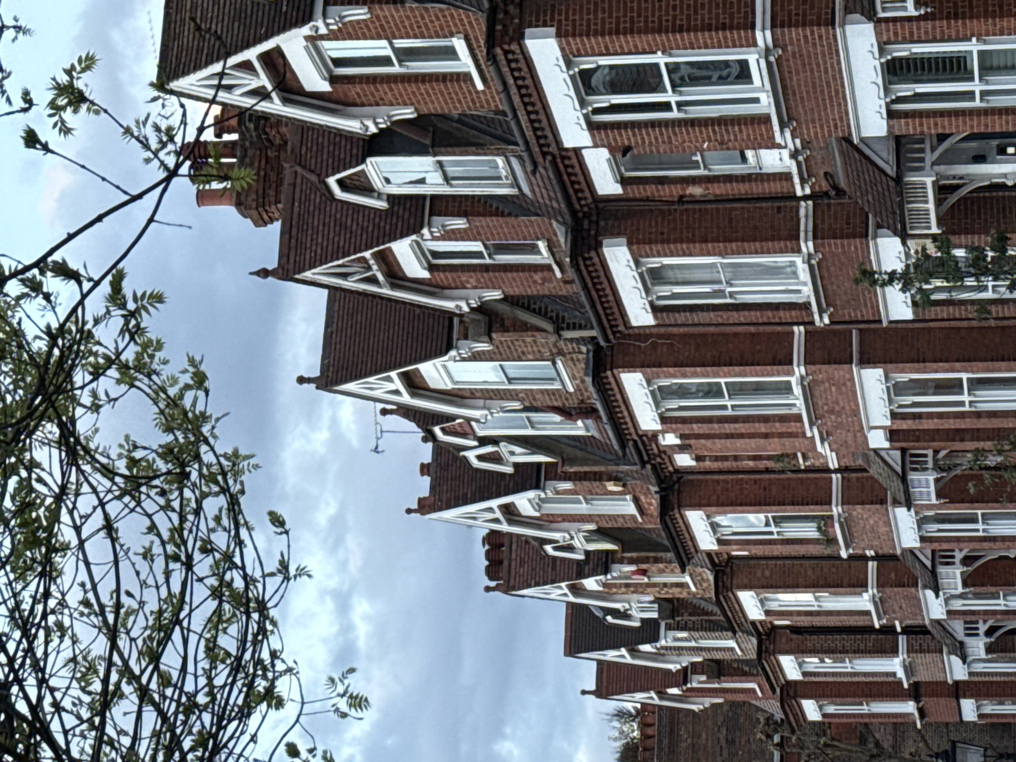 The image shows a row of red brick terraced houses in London, United Kingdom. Each house has a steeply pitched roof with white decorative bargeboards and small dormer windows. The facade consists of red brick with white window frames. A tree with bare branches is visible on the left side of the image, partially obscuring the view of the houses. The sky is overcast with grey clouds.