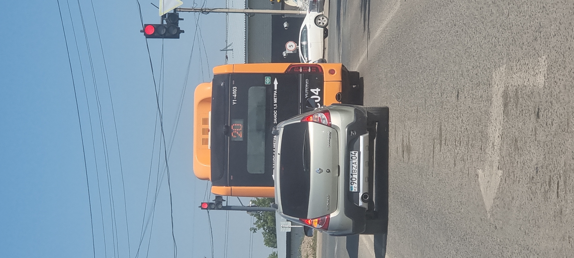 In Aktobe, Kazakhstan, on a clear day with a bright blue sky, a traffic scene unfolds. A silver Renault Sandero hatchback is stopped at a traffic light, facing away from the camera. Its license plate reads "KZ 901 BZA 04". Behind it, an orange and black Yutong bus is also stopped. The bus displays the number "20" in orange illuminated digits on its rear window, along with the designation "Y1-4003". A red traffic light is visible in the upper right portion of the frame. A white sedan is parked on the right side of the road. The scene is set on a paved road with lane markings. The surrounding environment suggests an urban or suburban setting with some foliage visible in the distance.