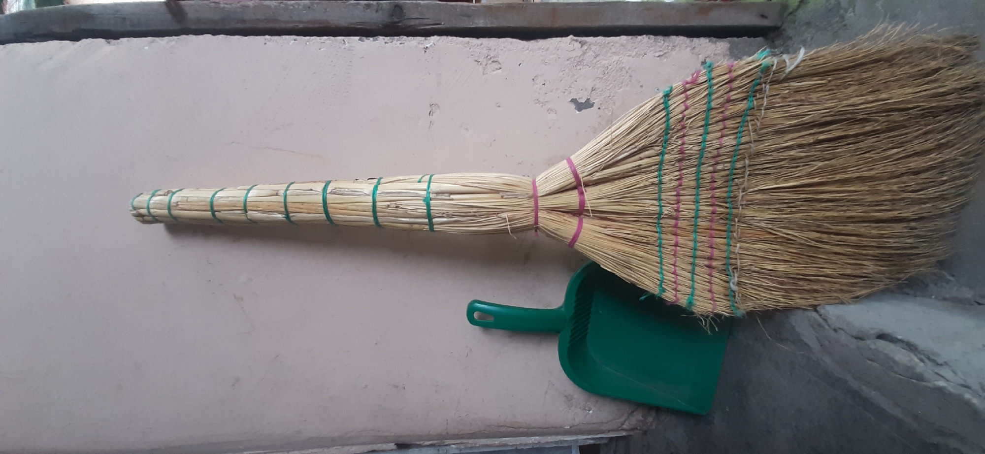 A traditional straw broom and a green plastic dustpan are positioned against a light beige, textured wall. The broom, tied with several green and pink rubber bands, stands upright and occupies the right half of the frame, its straw bristles fanned out at the bottom. The dustpan, with its handle facing left, rests on a gray concrete surface to the left of the broom. The floor appears cracked in places. The scene is indoors, with the wall possibly being an interior or exterior surface. The lighting is moderate, casting soft shadows. There are no people visible in the image.