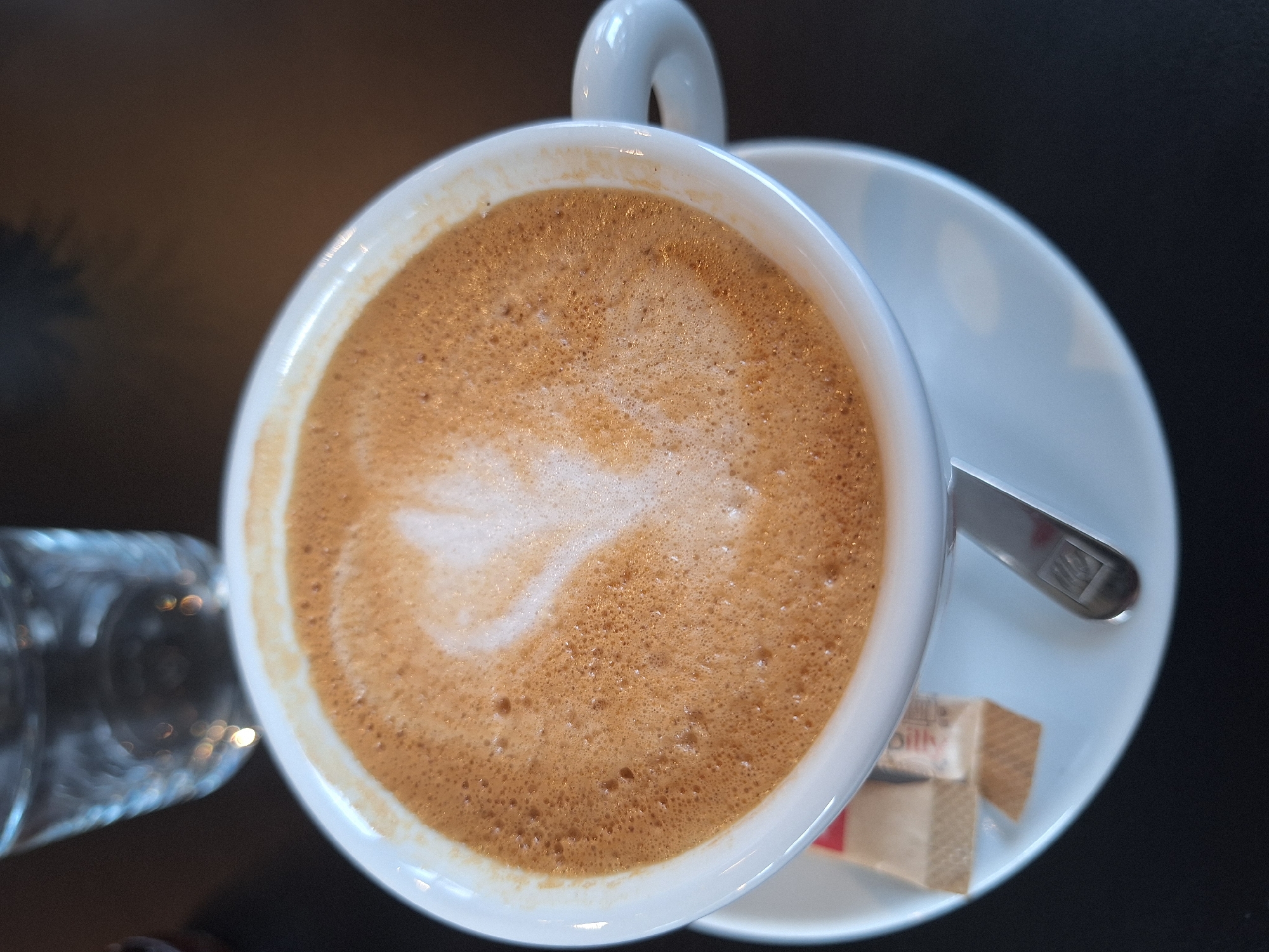 The image is a top-down view of a cup of coffee, likely a cappuccino or latte, with latte art resembling a leaf or feather, situated on a white saucer. A small spoon rests on the saucer to the right of the cup, next to a sugar packet. A glass, potentially containing water, is partially visible on the left side of the frame. The background is a dark, reflective surface, possibly a table.
The location is unknown, it is not possible to identify if the picture was taken in the city of Varna, Bulgaria. There are no discernible people or actions visible in the frame.