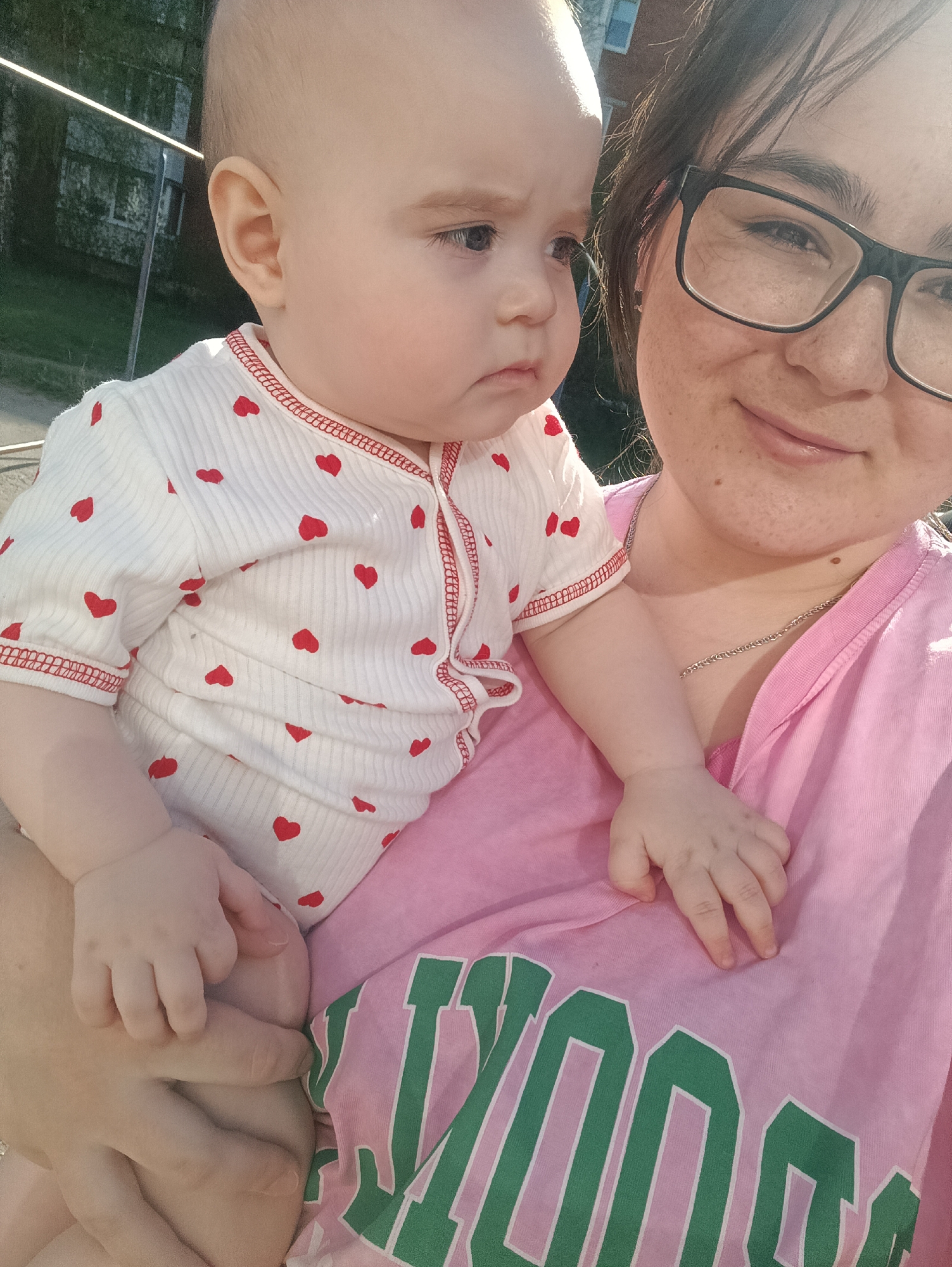The image shows a woman holding a baby. The woman has dark hair, glasses, and is wearing a pink shirt. The baby is wearing a white shirt with red hearts. They appear to be outdoors in a residential area. It is impossible to determine if the location is Noviy, Russia, as there are no contextual clues that would indicate the city's location.