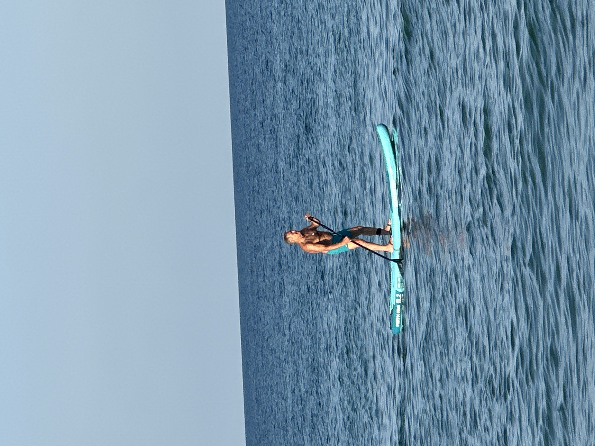 A man is paddleboarding on the ocean. He is standing on a light blue paddleboard and holding a paddle. He is wearing blue swim trunks and no shirt. The ocean is blue and there are small waves. The sky is clear and blue.