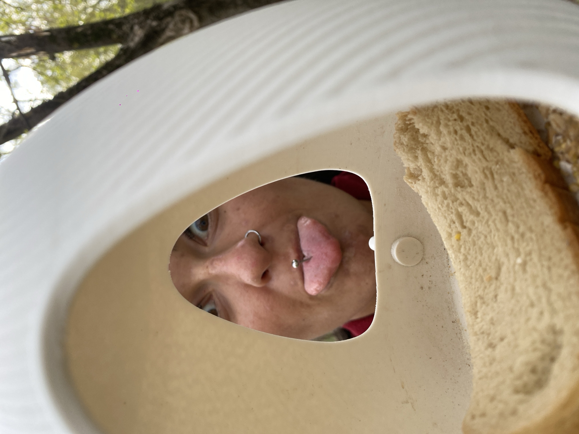 A person is looking down into a white container. Inside the container are slices of bread. The person has a nose ring and a tongue piercing. The background is blurred, but appears to include trees. Location information is not present in the image.