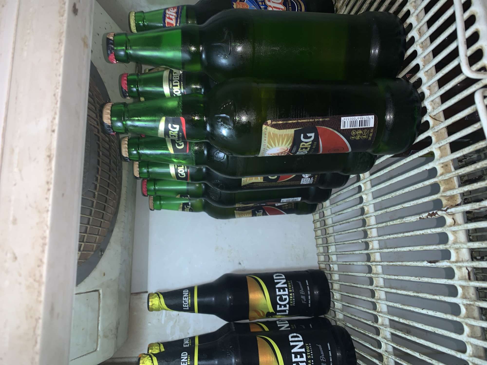 The image shows a shelf inside a refrigerator filled with several bottles of beer. On the left side of the shelf are four dark-colored bottles with yellow and black labels that read "LEGEND EXTRA STOUT PREMIUM QUALITY FULL BREWED". To the right of these are multiple green glass bottles of beer. Several of these green bottles have labels with text that appears to include "GOLDBERG PREMIUM LAGER" and "Life". The refrigerator shelf is made of white wire grating, and some of the bottles appear to be condensation-covered. The background inside the refrigerator is white, and a fan grille is visible at the top. The scene is located on the shelf of a refrigerator, within a residence in New Stadium, Nigeria.
