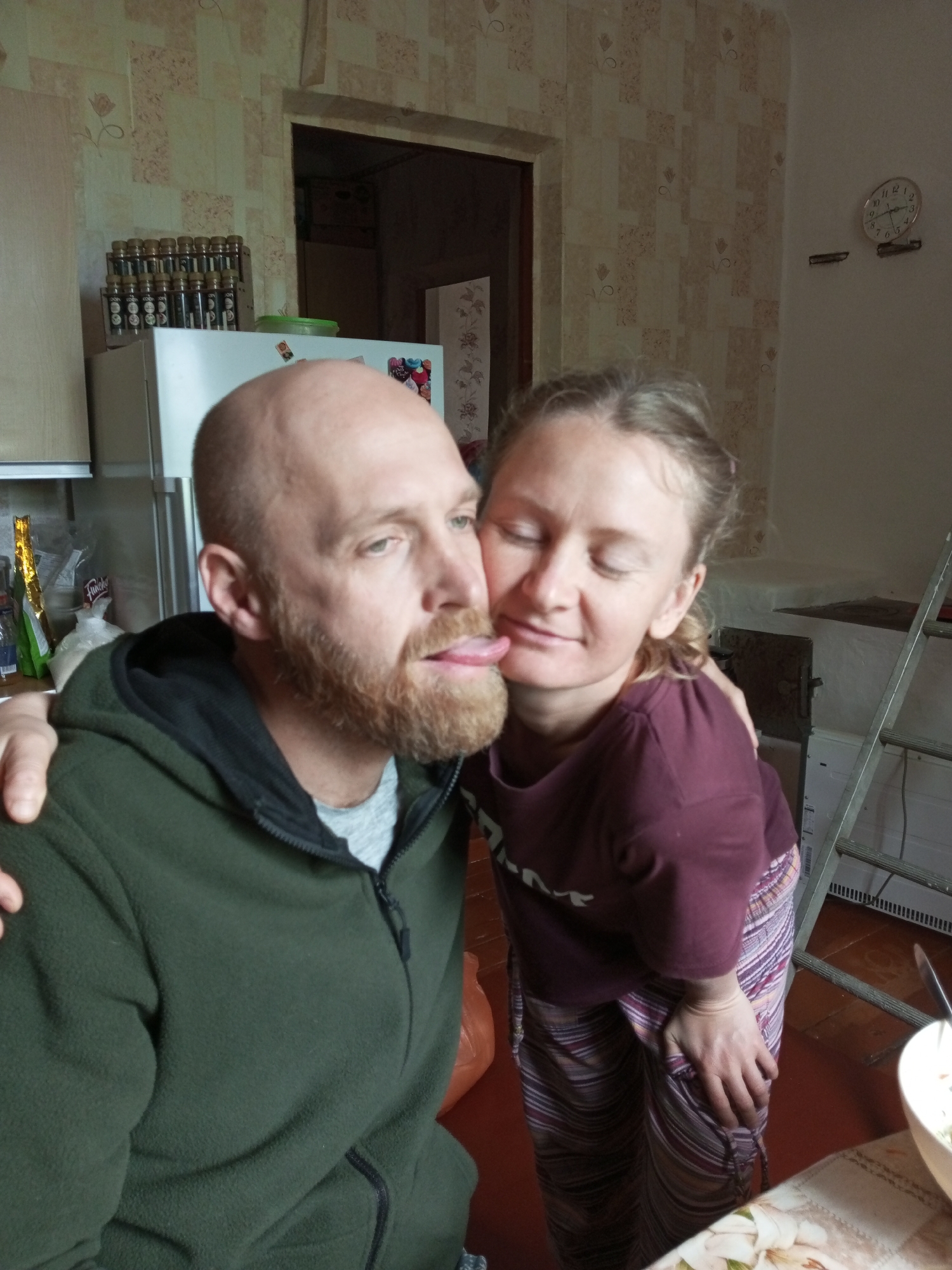 A medium shot photograph captures a couple in an indoor domestic setting, likely a kitchen. The man, on the left, is bald with a beard, wearing a green zip-up fleece jacket over a grey t-shirt. He is looking to the left with his tongue sticking out. The woman, on the right, has her eyes closed and is leaning in to kiss the man on the cheek. She has blonde hair tied back and is wearing a maroon t-shirt and striped pants. Her arm is around the man's shoulder.
In the background, a refrigerator is visible, with bottles of spices on top of it. Further back, a doorway leads to another room, and a clock hangs on the wall. To the right, a ladder leans against the wall, and a bowl of food sits on a table. The walls are covered in patterned wallpaper. The overall impression is of an informal, affectionate moment shared between the couple within their home.
