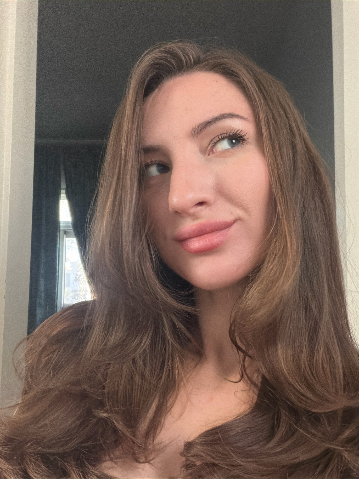 A medium shot shows a young woman with long, brown, layered hair looking to her left with a slight smile. Her eyes are blue, and she is wearing lip gloss. She is indoors, in what appears to be a bedroom, with dark curtains visible in the background behind a window. A white door frame is on the left side of the image. There is no indication of the city of Sharm El Sheikh, Egypt, in the image.