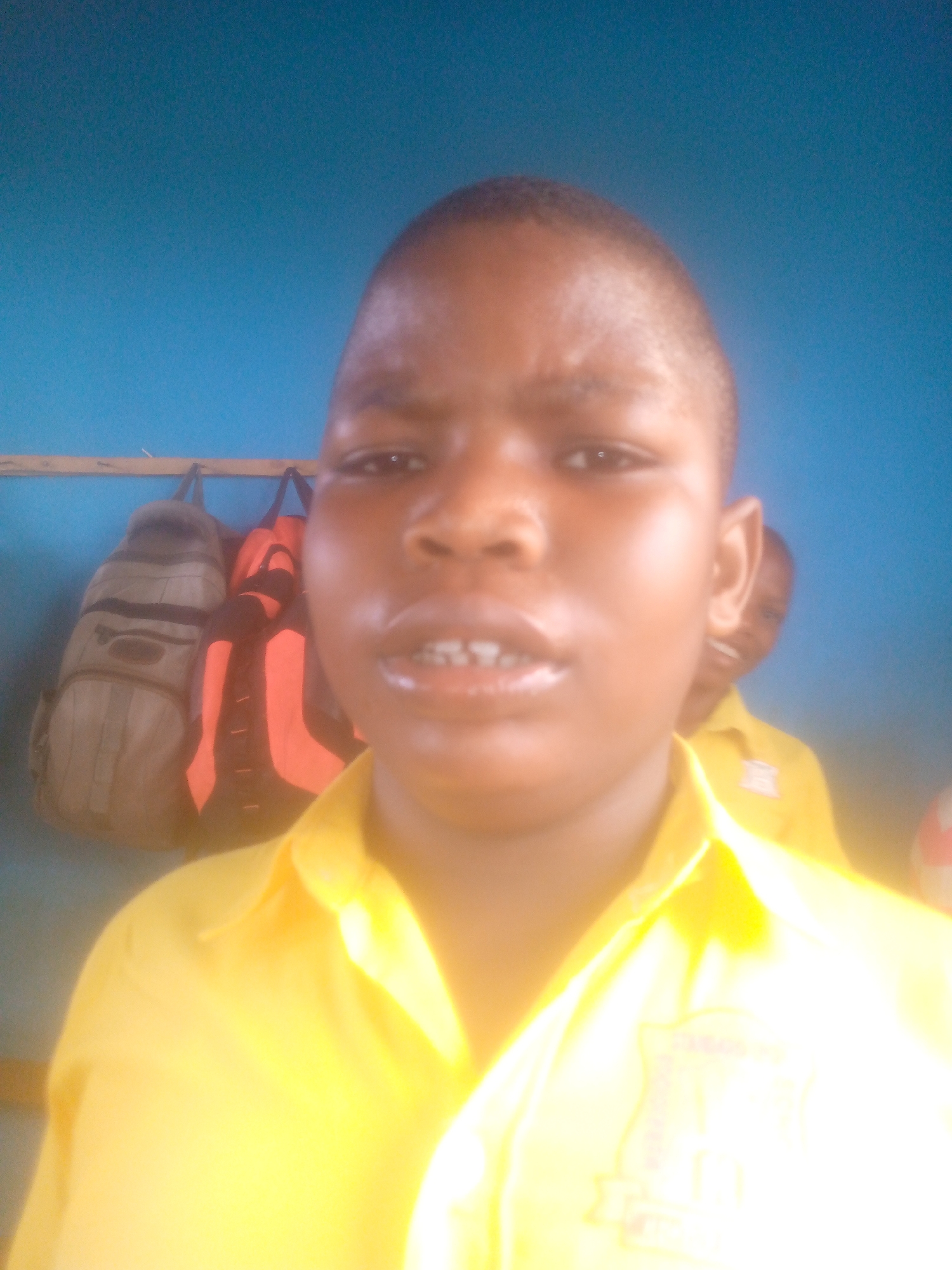 A young person is positioned close to the camera, wearing a yellow collared shirt, with other people and items visible in the background. Behind the subject, on a rail, are two bags, one tan and brown, and another red and black. Another young person is partially visible behind and to the right of the main subject, also wearing a yellow collared shirt. The background is a blue wall, possibly indicating an indoor location such as a classroom. The exact location within the city of Unknown, Unknown is not discernible.