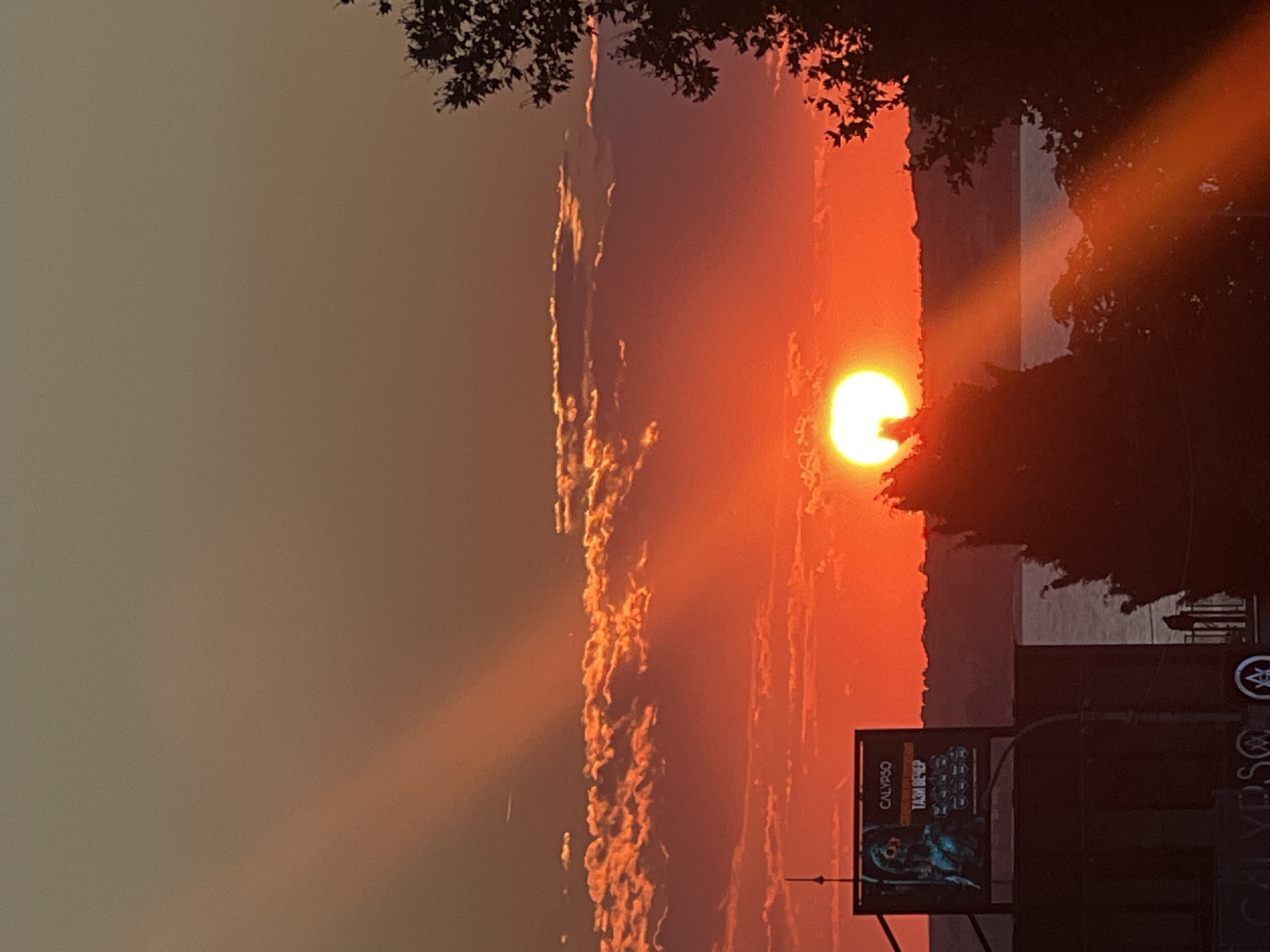 The image depicts a sunset over the sea in Sozopol, Bulgaria. The sun, a bright orange orb, is partially obscured by silhouetted trees and the dark shapes of buildings in the foreground. Rays of sunlight stream through the sky, illuminating scattered clouds tinged with orange and pink hues. A billboard featuring a figure and text in what appears to be Cyrillic script is visible on the left side of the frame. The water of the sea is a muted gray under the dramatic sky. The overall impression is one of a vibrant and atmospheric sunset in a coastal city.