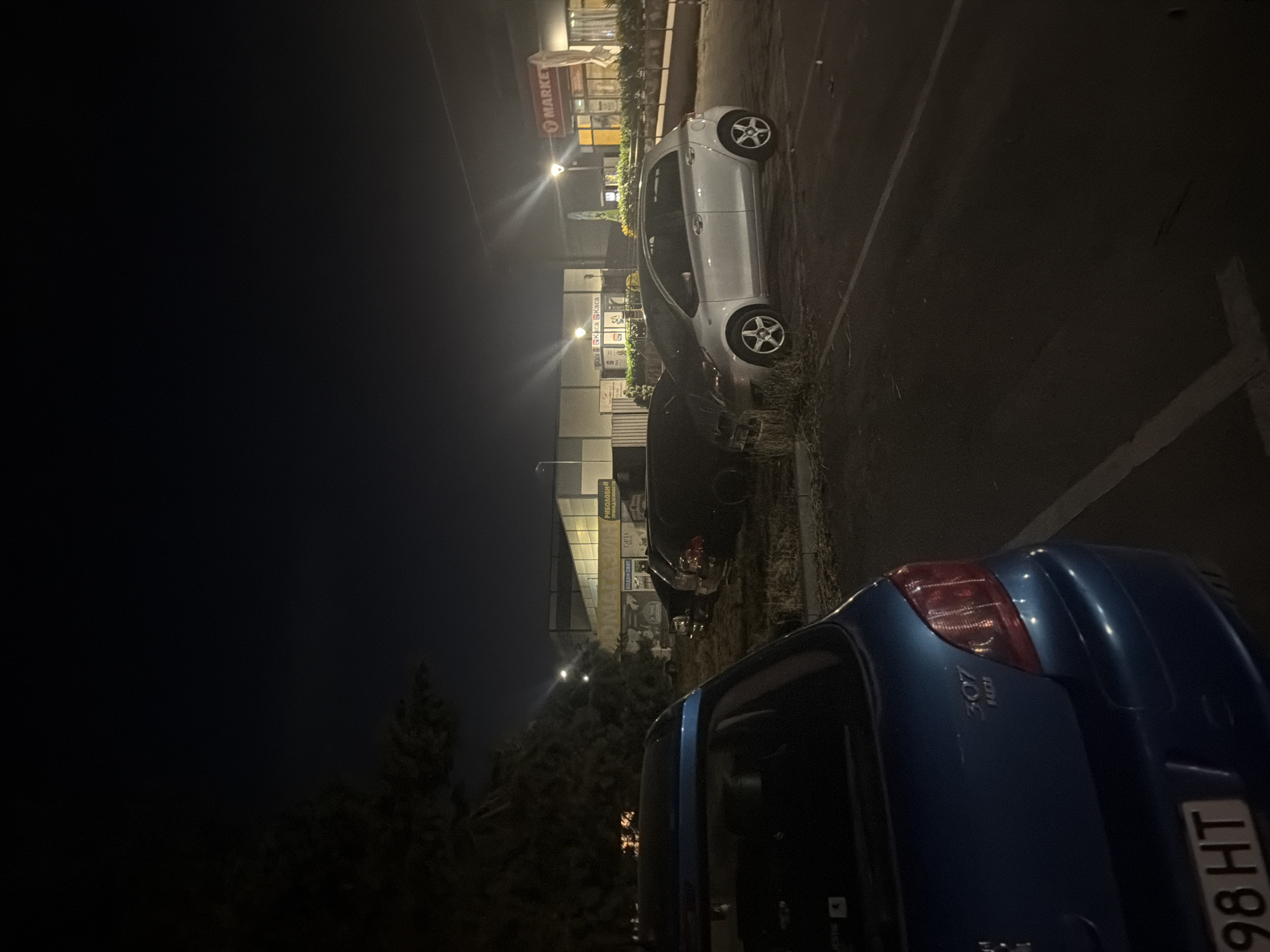 The image depicts a dimly lit outdoor parking lot at night. In the foreground, the rear of a blue Peugeot 307 HDI is visible, with its license plate reading "98 HT". To the right, a silver hatchback, possibly a Toyota Yaris, is parked. In the background, a commercial building with signage including "MARKET" is illuminated. The building appears to be a retail or commercial establishment. Several other dark-colored vehicles are parked in the lot, some partially obscured by foliage. The ground is asphalt, with visible parking space lines. The overall scene suggests a typical urban parking area at night, potentially near a shopping center or market. The location is identified as Plovdiv, Bulgaria.