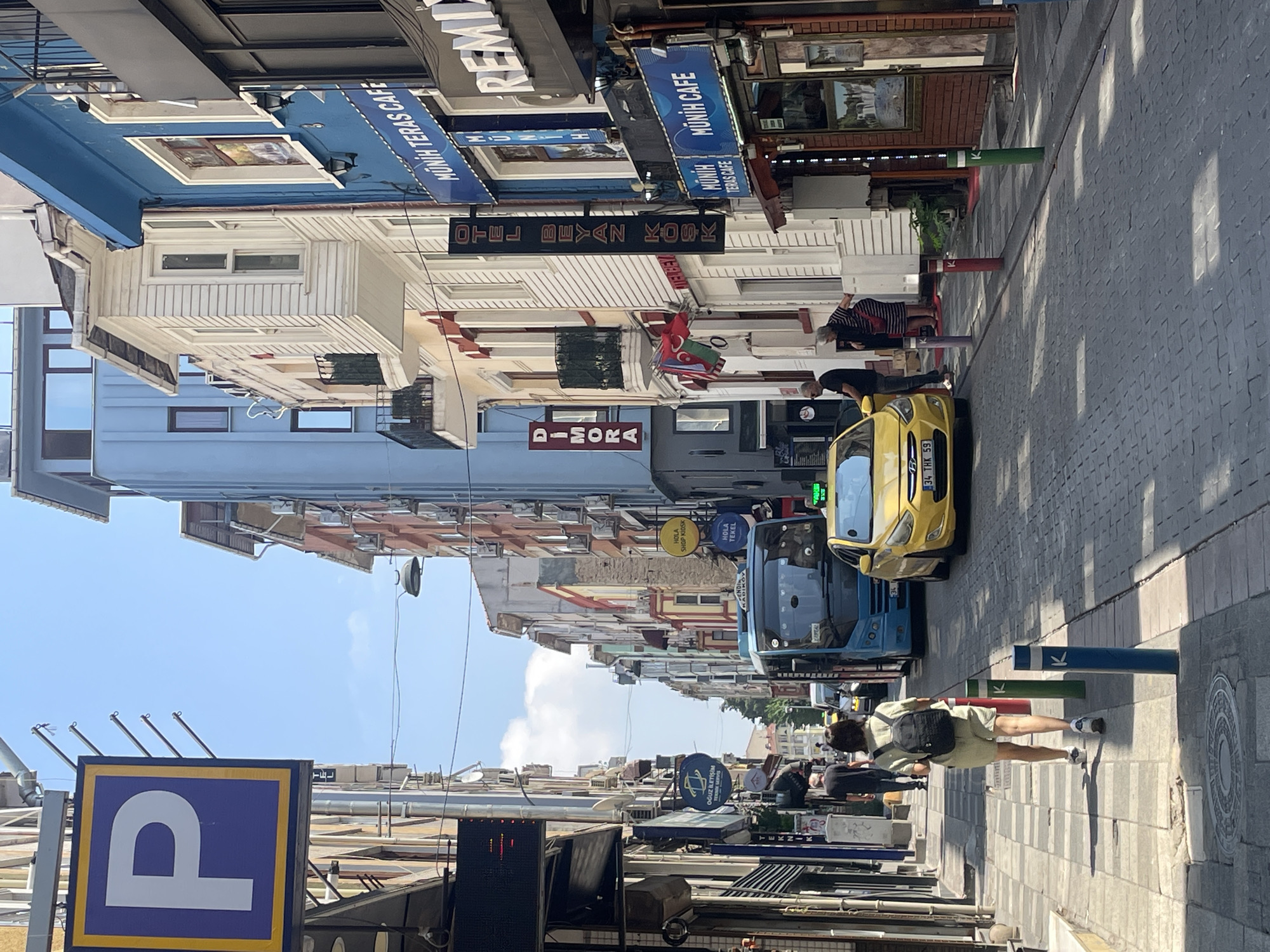 In Kadıköy, Turkey, on a sunny day, a narrow street is lined with multi-story buildings of varying architectural styles and colors. A blue bus and a yellow taxi are parked on the cobblestone street. A woman with a backpack is walking away from the camera on the sidewalk, which is also paved with cobblestones. Several signs are visible on the buildings, including "P" parking sign, "DİMORA," "OTEL BEYAZ KÖSK," and "MÜNİH TERAS CAFE." Some people are standing on the sidewalk near the taxi and cafe. The sky is clear with a few clouds.