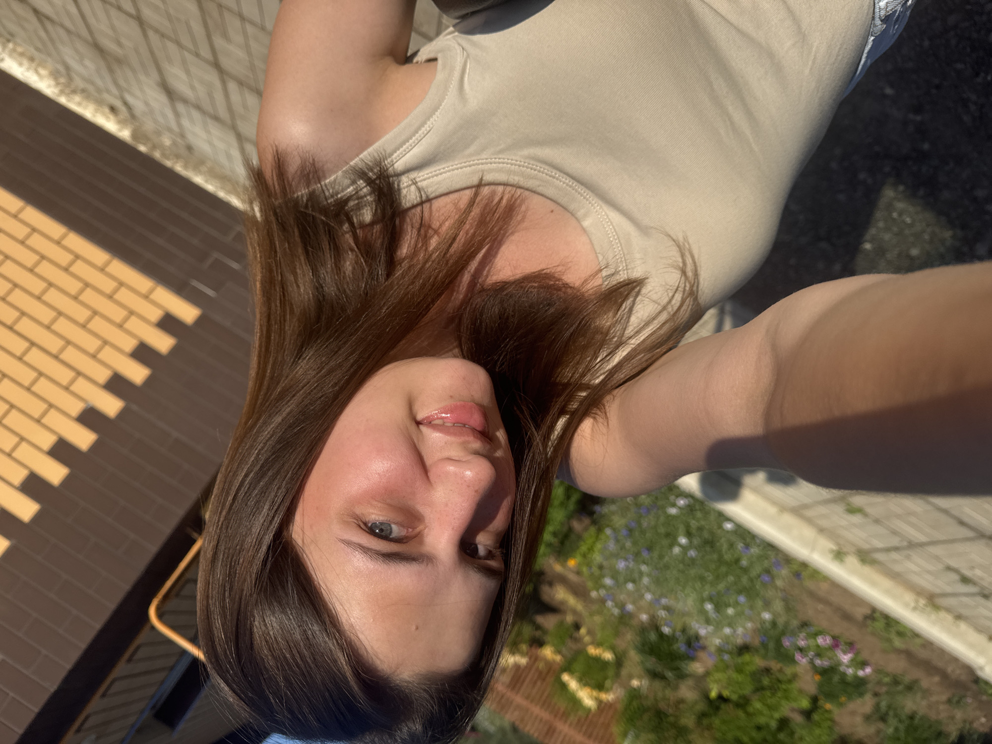 A young woman with long brown hair and blue eyes smiles and looks at the camera. She is wearing a beige tank top. The background is blurry, showing a garden with flowers and a building with a brick pattern. The image appears to be taken outdoors in Vereshchagino, Russia, possibly during a sunny day.