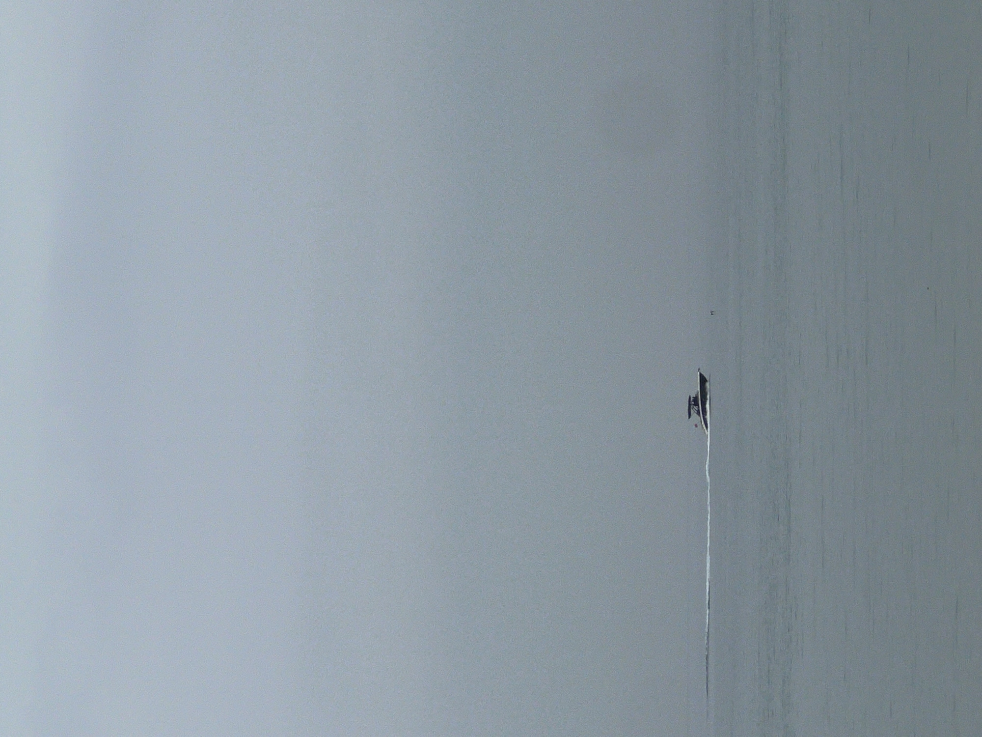The image captures a minimalist scene of a solitary boat navigating a vast expanse of calm water under an overcast sky. The dominant colors are various shades of grey, blending the sky and water into an almost seamless, muted backdrop, making the horizon nearly indistinct.
A small, dark-colored boat is positioned in the upper right portion of the frame, moving towards the upper edge. A long, distinct white wake trails behind the boat, indicating its active movement through the water. Due to the distance and resolution, specific details of the boat's design or any occupants are not discernible.
The water surface appears smooth and calm, with subtle textural variations, particularly noticeable on the right side, suggesting gentle ripples or reflections. The sky is uniformly light grey, denoting an overcast day. There are no clear indications of the time of day beyond it being daylight, and the weather appears calm and possibly hazy or foggy in the distance.
The setting is consistent with a coastal environment, such as the location provided, M'Diq, Morocco, which is situated on the Mediterranean Sea. No people or visible text are clearly identifiable in the image. The overall impression is one of tranquility and solitude.