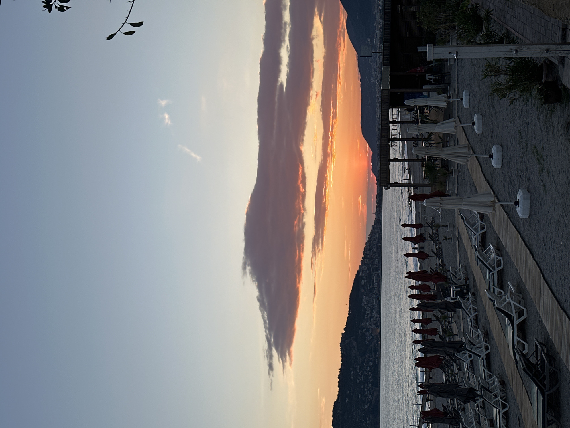 The image depicts a beach scene, likely at sunset, in an unspecified location. A body of water, probably the sea, is centrally located and extends to the base of a mountain range in the background. The mountain has buildings along its slope. The sky is filled with clouds that transition from a lighter hue to orange and pink due to the setting sun. The beach has rows of lounge chairs and umbrellas. The image is taken from a high angle, and a branch with leaves is present in the upper left corner. The image does not contain the city of Alanya, Turkey.