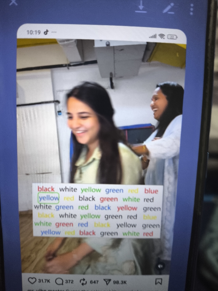The image displays a photograph of a vertically held mobile phone screen. The screen exhibits an application interface, likely TikTok, at 10:19, showing Wi-Fi connectivity and 72% battery. The primary content on the phone screen is a video or image featuring two women in an indoor setting.
The woman in the foreground, positioned left, has long, dark hair and wears a light green or pale yellow collared shirt. She is captured smiling broadly, looking down and to her right. Her hands are partially visible at the bottom of the frame, resting below a text overlay. The woman in the background, positioned right, also has dark hair and is smiling broadly while looking towards the first woman. She is wearing a light-colored, possibly white, long-sleeved garment with visible textured or embroidered details and has her arms crossed.
The background visible within the phone content shows white walls and exposed, light-colored rectangular ductwork or beams on the ceiling. A dark horizontal line is present in the upper background, and a small blue object is visible on the left wall. A bright yellow element is discernible in the upper right.
Superimposed over the lower half of the women is a multi-line text overlay presenting a color-word challenge, similar to the Stroop Effect. Words like "black," "white," "yellow," "green," "red," and "blue" are displayed in varying ink colors, sometimes matching the word (e.g., "black" in black ink) and sometimes conflicting (e.g., "yellow" in green ink, outlined in green). Below this, application UI elements include a heart icon with "31.7K," a comment bubble with "372," a share icon with "647," and a bookmark icon with "98.3K." A partial caption "mr. vibe master Guess the color not the word wh" is visible at the very bottom.
There are no discernible visual elements within the image that allow for the identification of a specific location within Nicosia, Northern Cyprus, or Cyprus. The environment depicted within the phone's content is a generic indoor space.