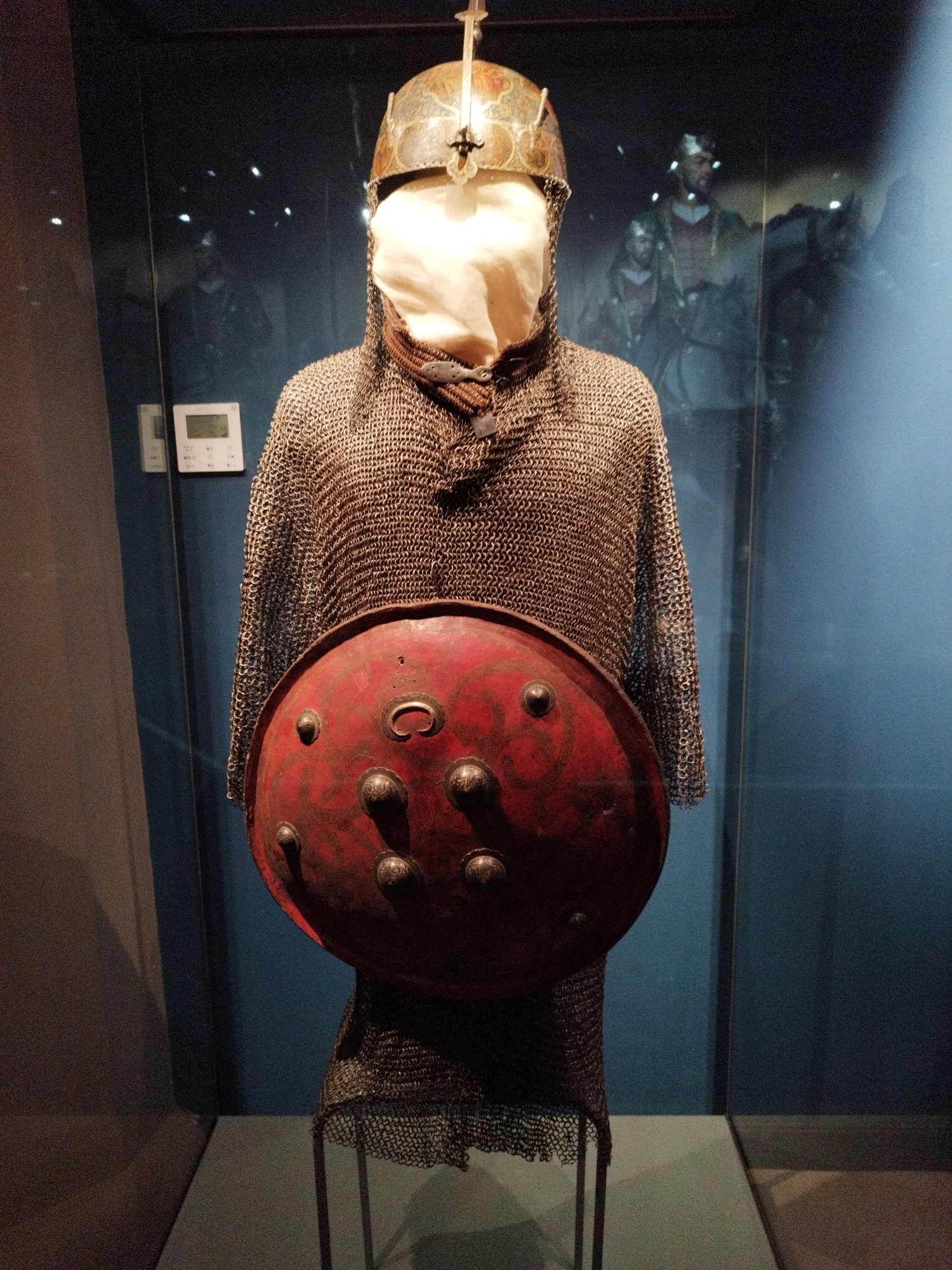 This is a full-frame, eye-level shot of a suit of armor displayed in a glass-enclosed exhibit. The armor is worn by a mannequin and consists of a metal helmet with ornate detailing, a chainmail shirt that extends down to the knees, and a large, round, red shield with decorative silver studs and a central ornate boss. The shield is held in front of the mannequin's torso. The background is dark blue and slightly out of focus, with reflections of other figures, possibly other exhibits or museum visitors, visible on the glass. To the left of the armor, a white digital display panel is mounted on the wall. The exhibit appears to be indoors, likely in a museum setting.