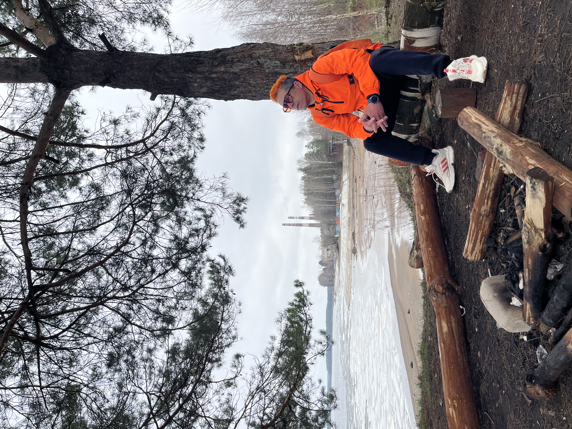 A person with orange hair and glasses, wearing an orange jacket and dark pants, is sitting on a wooden structure near a beach. The location is identified as Chaikovsky, Russia based on the two chimney stacks visible in the background. The beach appears to be covered in ice or snow, with water in the distance. A large tree is positioned to the left of the person, with its branches extending across the top of the frame. To the right, remnants of a campfire are visible with charred wood and ash. The sky is overcast.