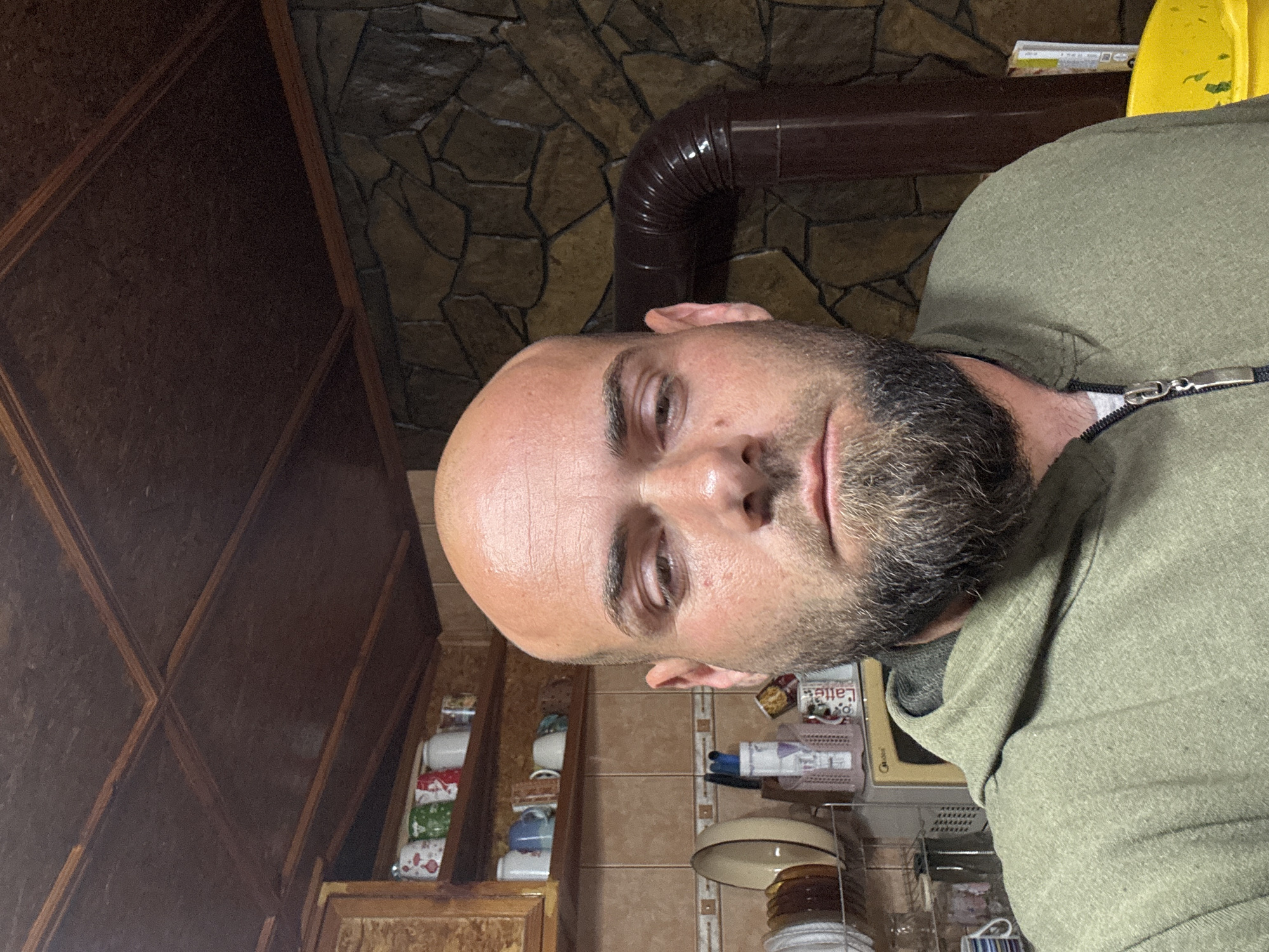 A rotated, close-up, eye-level shot of a balding man with a beard, wearing a green hooded shirt with a silver zipper, positioned in an indoor environment. Behind him are kitchen cabinets and utensils, a stone wall, and a brown pipe. Location is Sarnevo, Bulgaria.